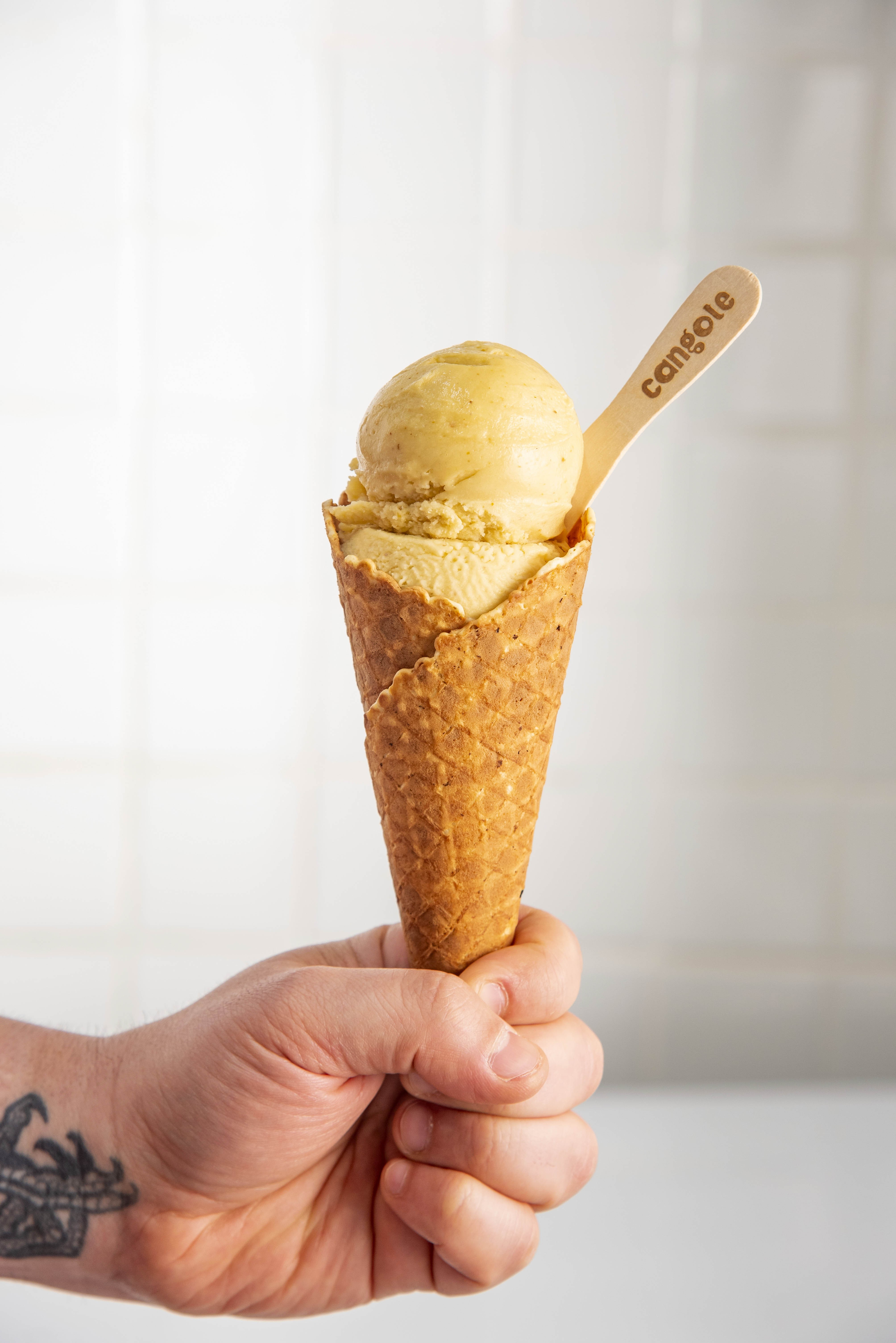 M&atilde;o masculina com tatuagem no pulso segurando uma casquinha de sorvete crocante com duas bolas de sorvete amarelo claro e uma colher de madeira com a palavra cangote gravada, em fundo branco