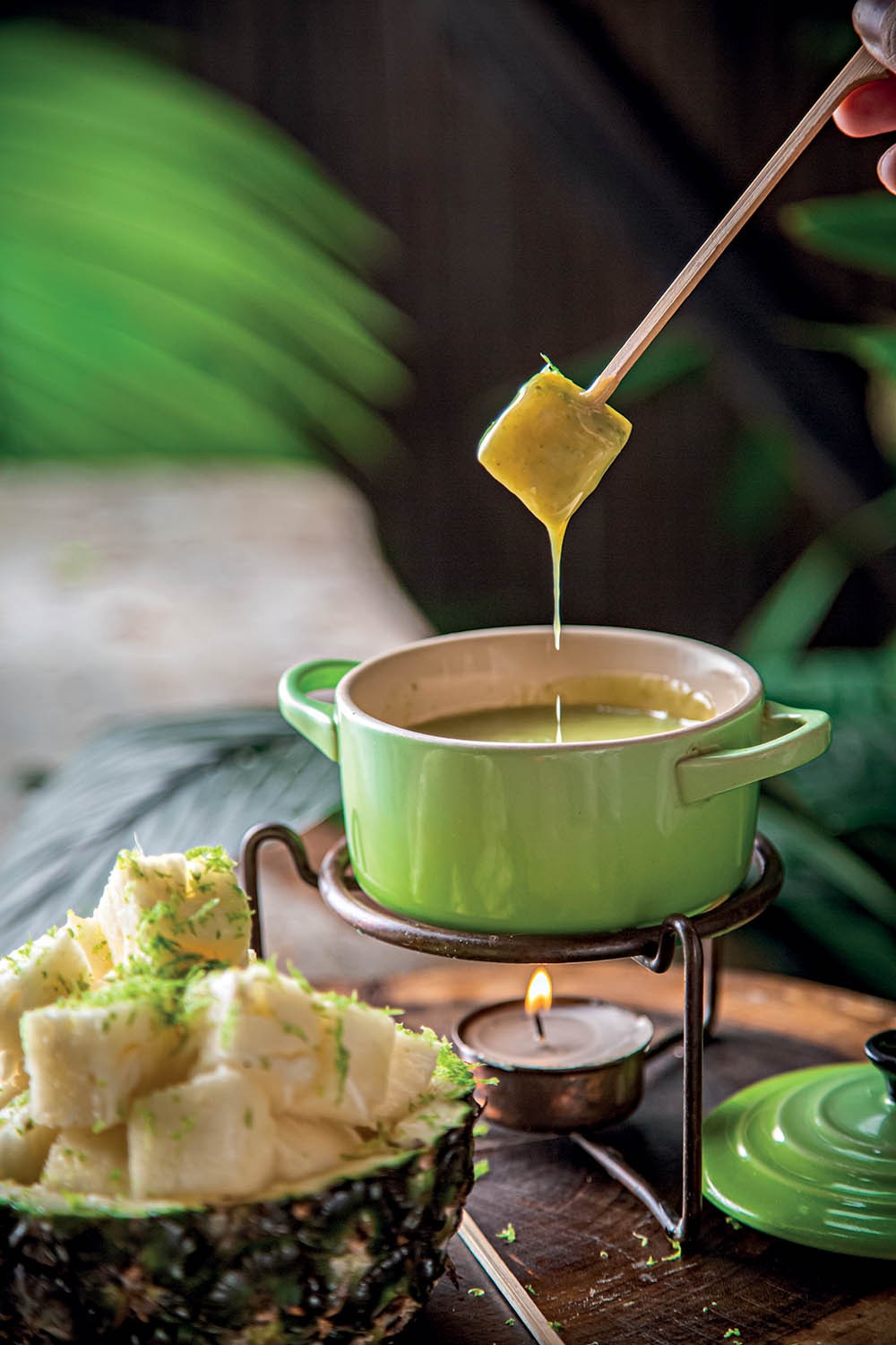 Uma m&atilde;o segura um espeto de bambu com um peda&ccedil;o de abacaxi mergulhado em um fondue verde claro, que escorre da fruta. O fondue est&aacute; em uma panela verde sobre um rechaud com uma vela acesa. Ao lado, uma metade de abacaxi com peda&ccedil;os da fruta picados e raspas de lim&atilde;o. Folhas verdes desfocadas ao fundo