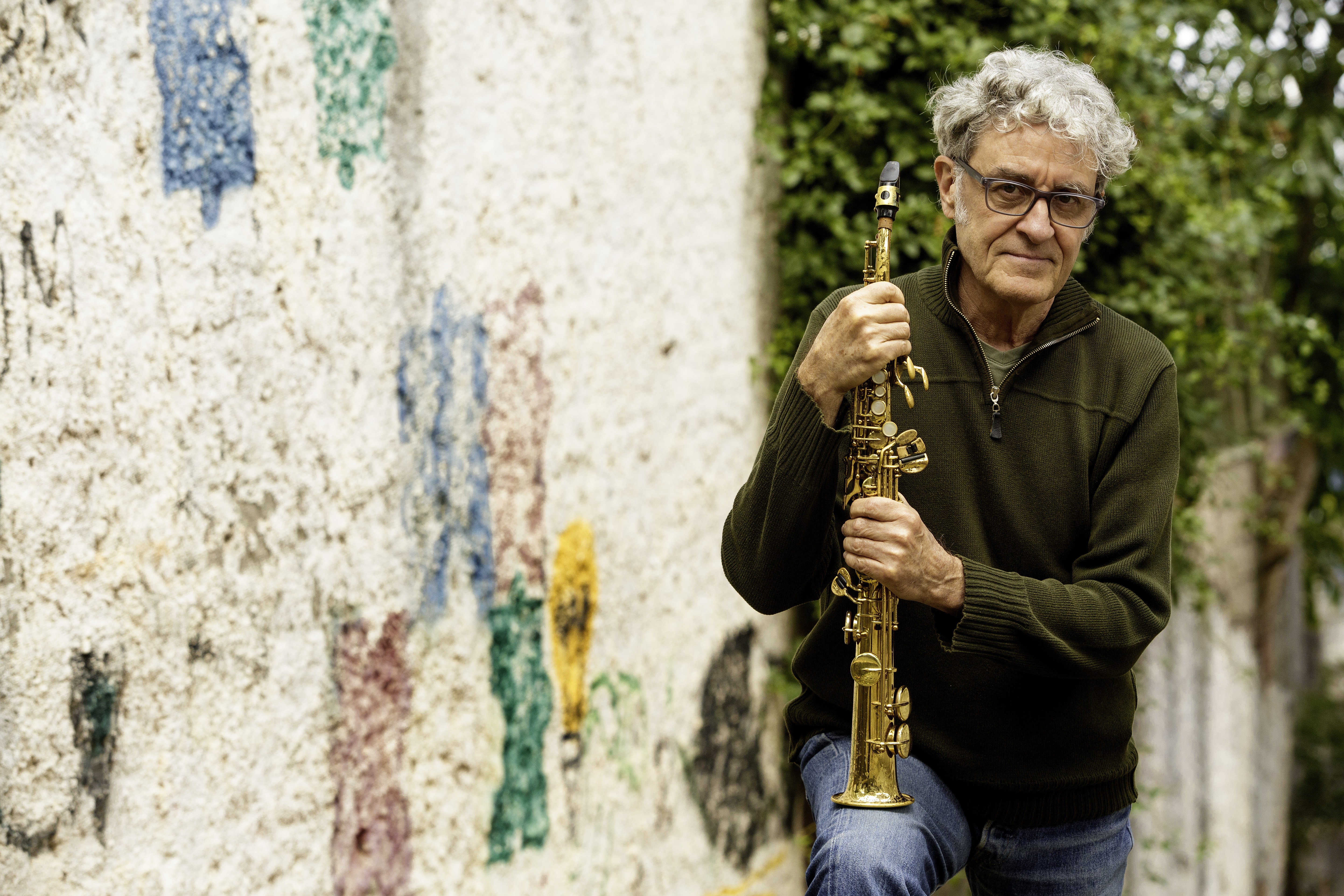 Homem de cabelos grisalhos e &oacute;culos, vestindo su&eacute;ter verde-escuro e jeans, segurando um saxofone dourado. Ele est&aacute; em frente a um muro claro com manchas coloridas, e folhagens verdes ao fundo