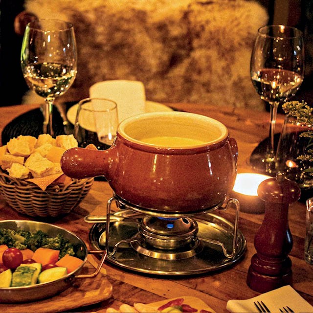 Uma panela de fondue de queijo borbulhante sobre um fogareiro, acompanhada por uma cesta de p&atilde;es, uma tigela de vegetais e ta&ccedil;as de vinho branco, em uma mesa de madeira com luz de velas