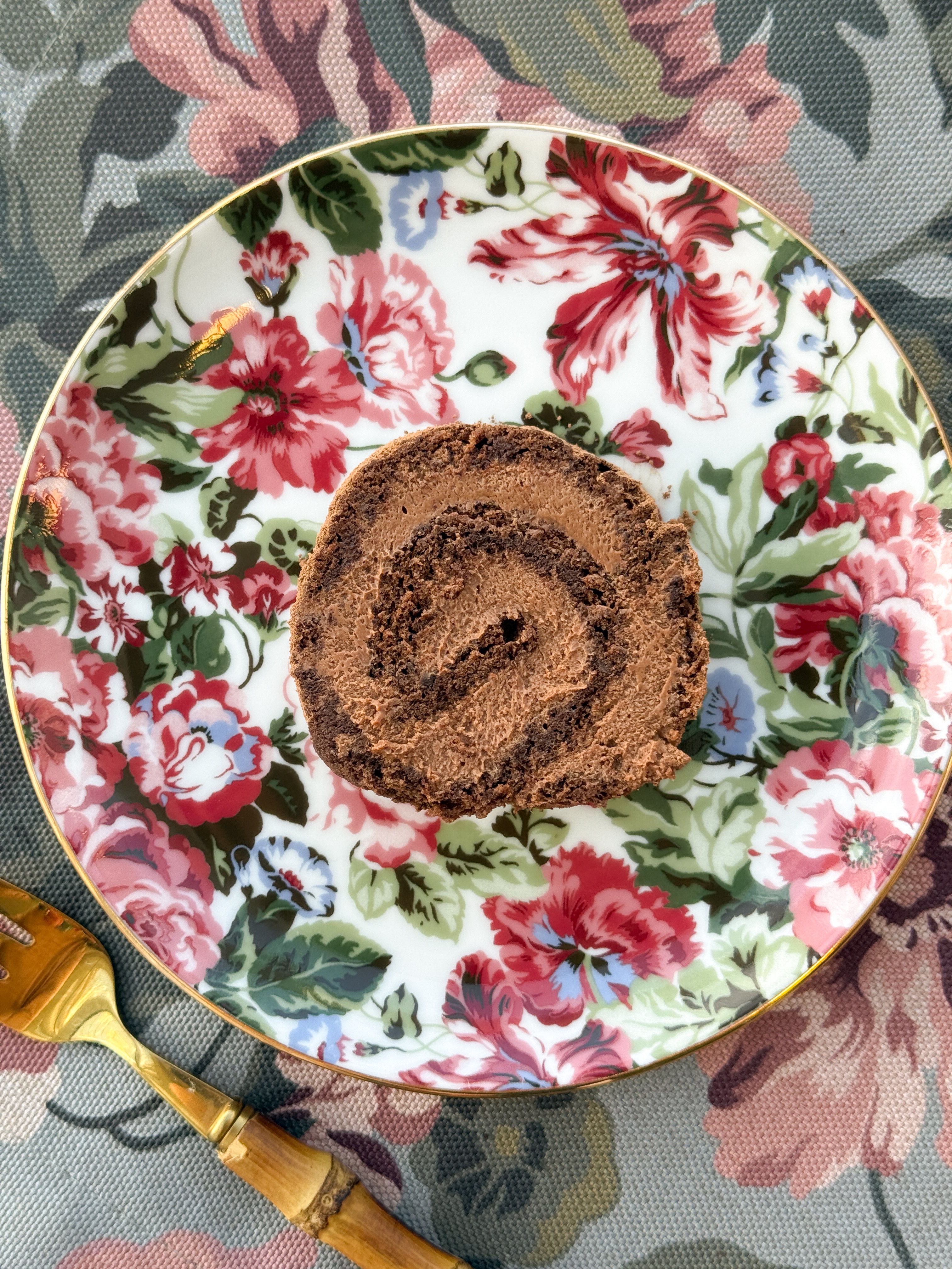 Fatia de rocambole de chocolate com recheio cremoso, servida em prato branco com estampa floral colorida e borda dourada, sobre toalha de mesa com padr&atilde;o de flores