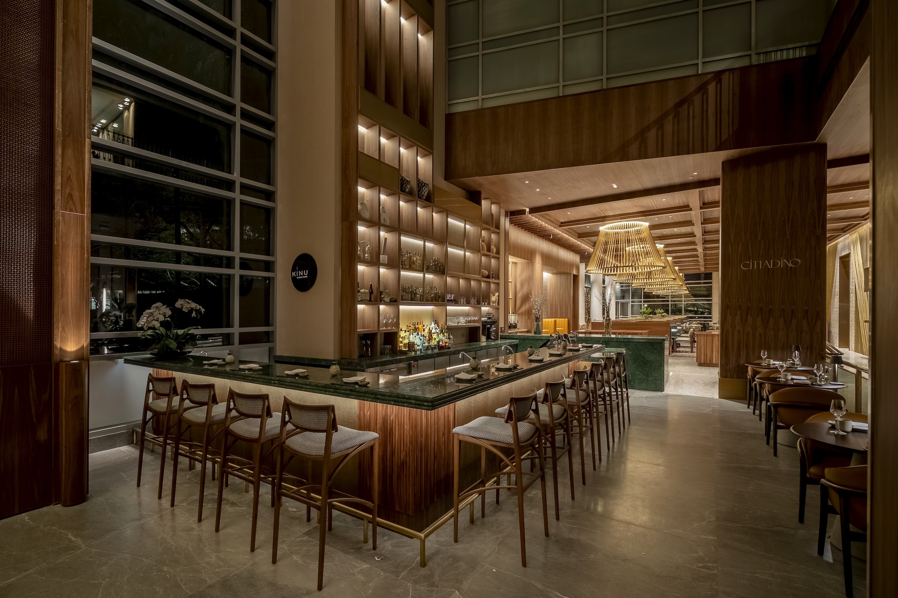 Interior de um restaurante moderno com balc&atilde;o de m&aacute;rmore verde e banquetas de madeira, iluminado por luzes quentes. Ao fundo, prateleiras com garrafas e um grande lustre de palha. Mesas de jantar com cadeiras escuras e janelas amplas completam o ambiente sofisticado