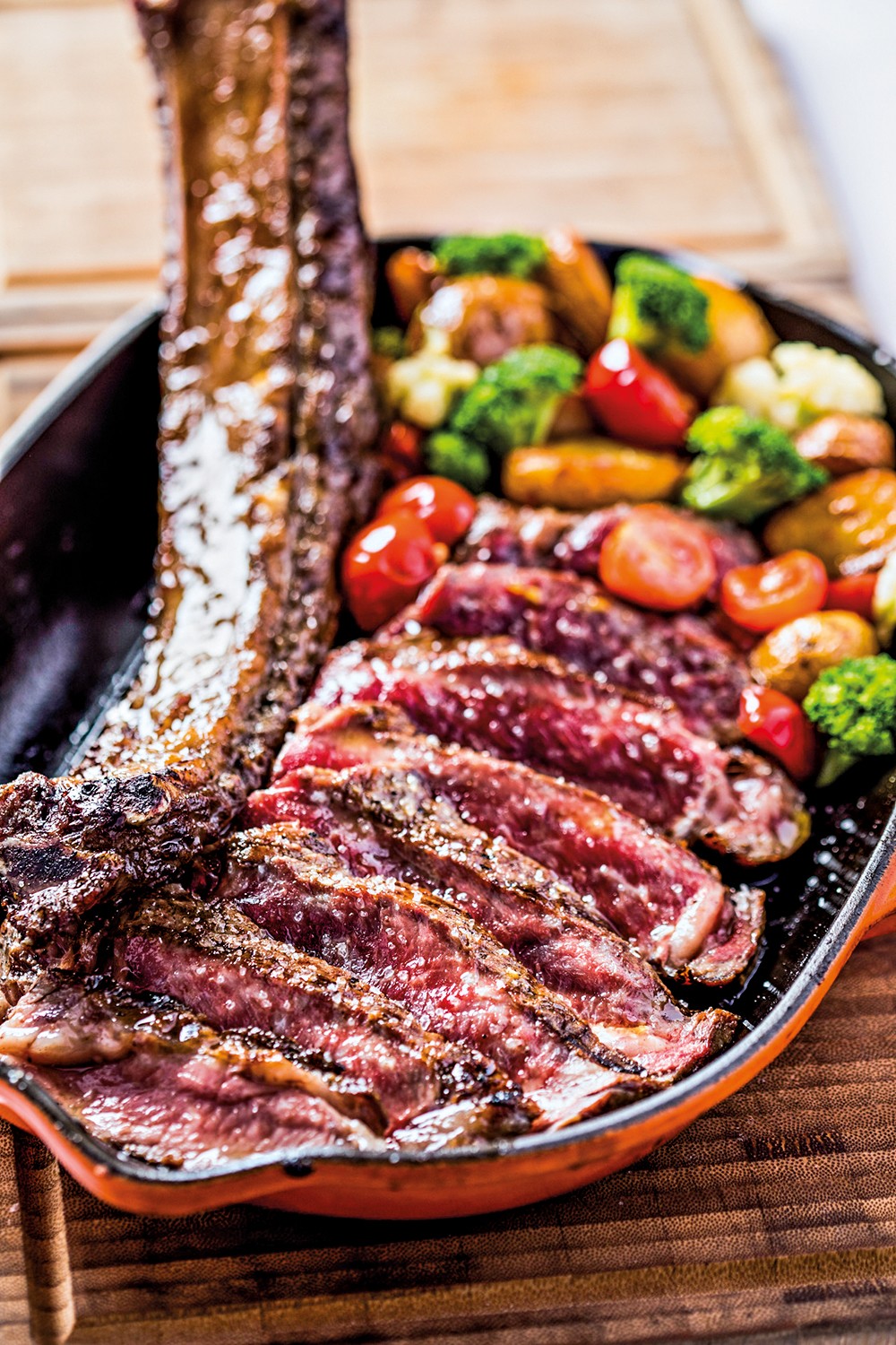 Bife tomahawk fatiado com osso, sal grosso e pimenta, acompanhado de br&oacute;colis, couve-flor, batatas e tomates cereja, servido em uma frigideira de ferro fundido laranja sobre uma t&aacute;bua de madeira