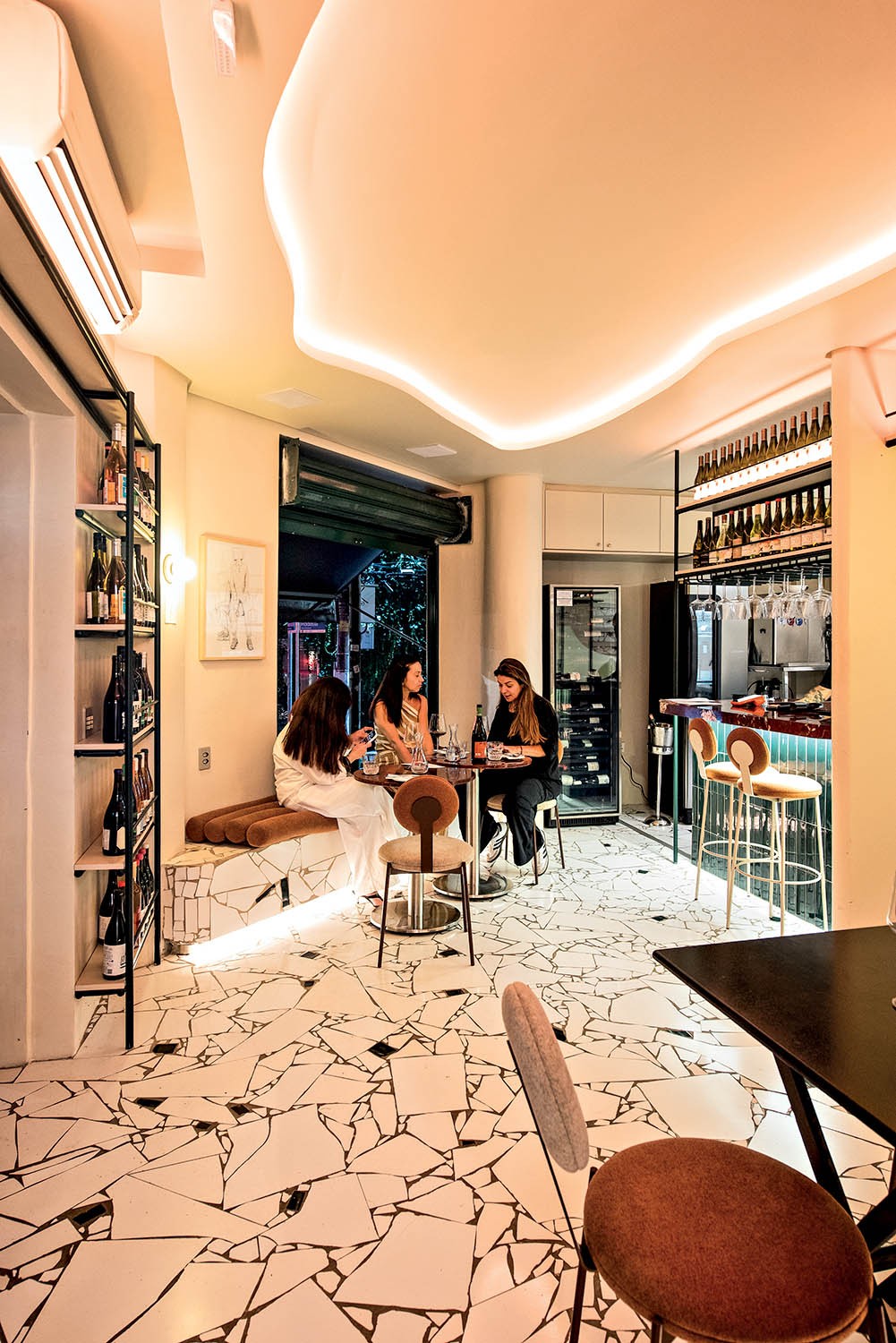 Tr&ecirc;s mulheres conversam em um bar com ilumina&ccedil;&atilde;o quente e teto ondulado. Duas sentadas em um banco estofado marrom, uma de blusa branca e outra de blusa preta, e uma terceira de blusa preta sentada em uma cadeira marrom. Elas est&atilde;o em uma mesa redonda com garrafas de vinho e ta&ccedil;as. O ch&atilde;o tem um padr&atilde;o de cacos brancos com rejunte escuro. &Agrave; esquerda, uma estante preta com garrafas de vinho. Ao fundo, um balc&atilde;o com garrafas e ta&ccedil;as, e uma adega climatizada