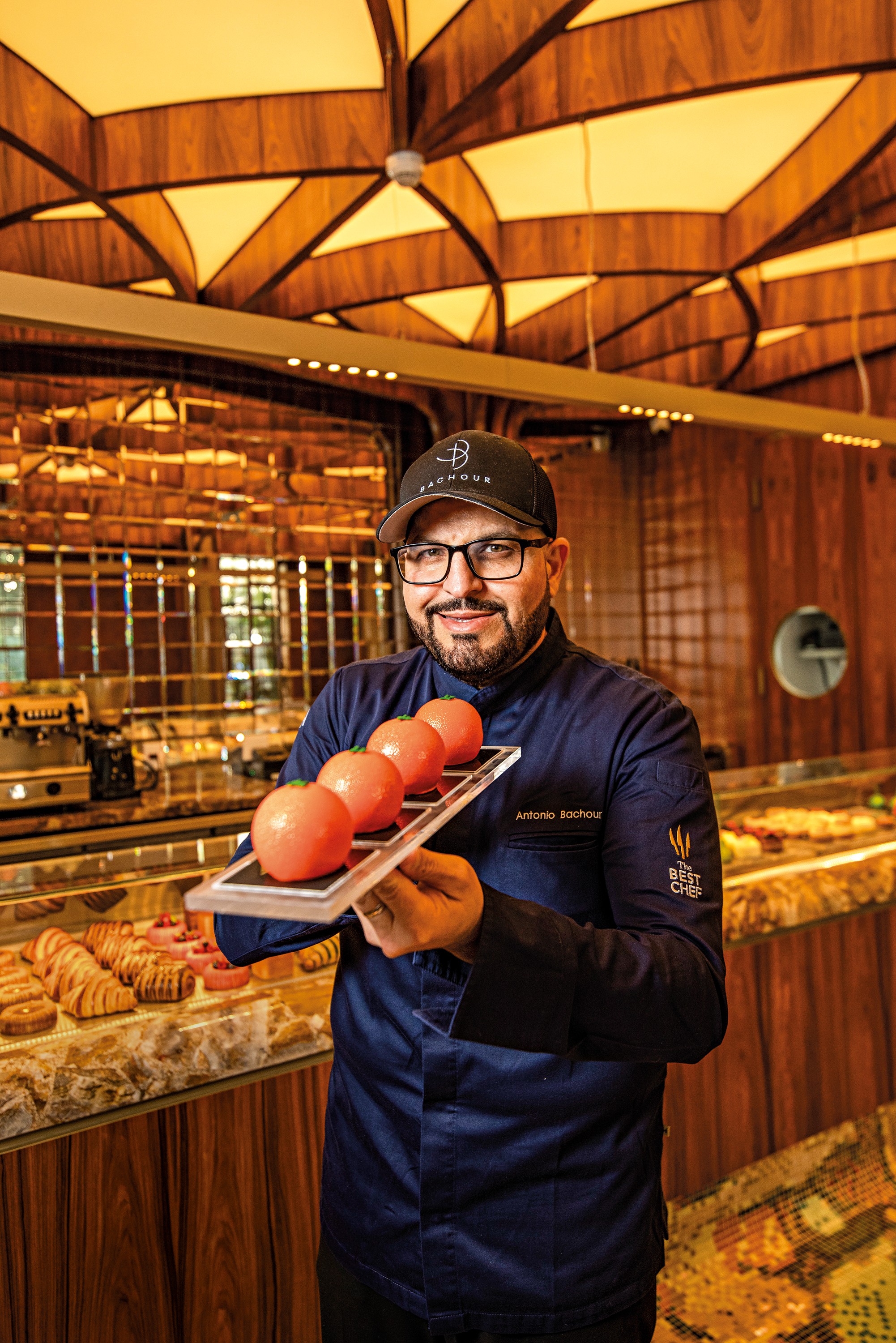 Homem de barba e &oacute;culos, vestindo uniforme de chef azul-marinho e bon&eacute; preto, sorri enquanto segura uma bandeja com quatro doces redondos cor de p&ecirc;ssego. Ao fundo, uma confeitaria com balc&otilde;es de doces e teto de madeira iluminado