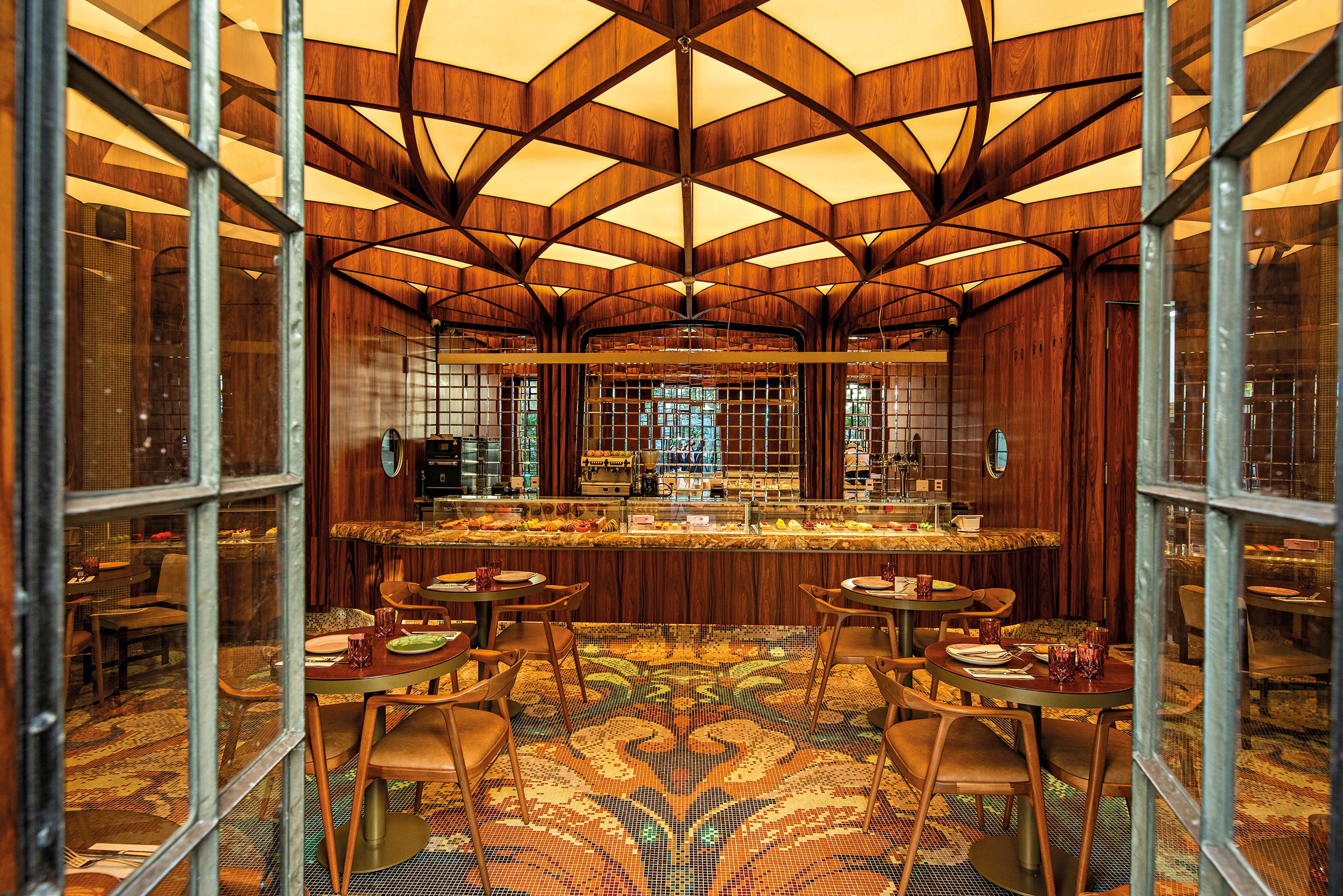 Interior de um restaurante luxuoso com teto abobadado de madeira e ilumina&ccedil;&atilde;o amarela suave. Ao centro, um balc&atilde;o de buffet com alimentos. Mesas redondas de madeira com cadeiras de couro marrom est&atilde;o dispostas sobre um tapete estampado com motivos de pav&atilde;o. As paredes s&atilde;o de madeira escura, e grandes janelas de vidro emolduram a cena