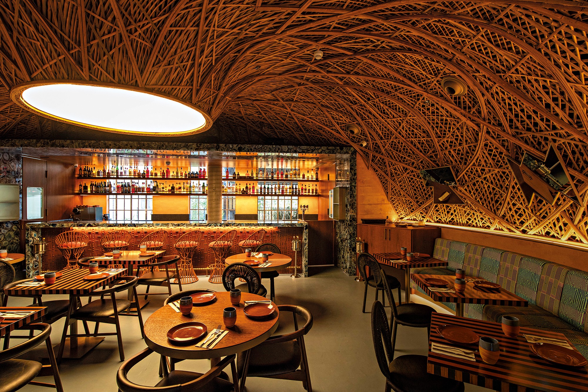 Interior de um bar ou restaurante com teto abobadado de madeira entrela&ccedil;ada. &Agrave; esquerda, um balc&atilde;o com garrafas iluminadas e banquetas de vime. Mesas de madeira com pratos e talheres est&atilde;o dispostas pelo sal&atilde;o, algumas com cadeiras escuras e outras com um longo sof&aacute; estampado. A ilumina&ccedil;&atilde;o &eacute; suave, criando um ambiente acolhedor