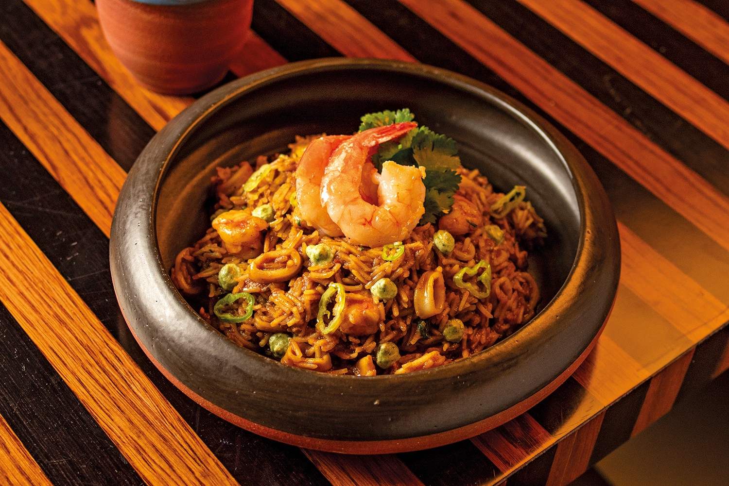 Arroz com frutos do mar, camar&otilde;es, lulas, ervilhas e piment&otilde;es verdes fatiados, servido em uma tigela de cer&acirc;mica escura sobre uma mesa de madeira com listras diagonais marrons e laranjas