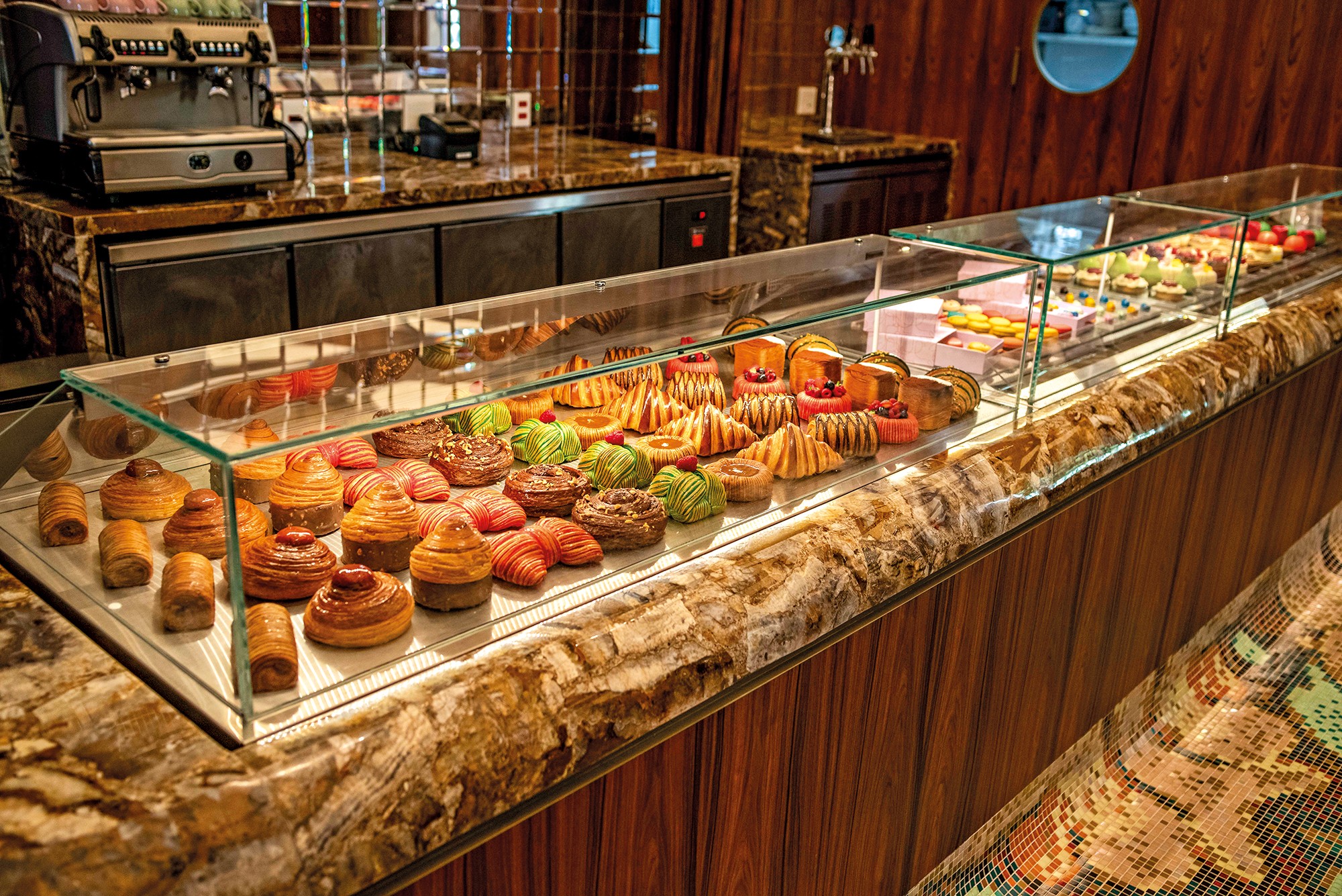 Exibi&ccedil;&atilde;o de doces e p&atilde;es em uma vitrine de vidro iluminada, com uma variedade de croissants, bolos e tortas coloridas. Ao fundo, uma m&aacute;quina de caf&eacute; expresso e balc&otilde;es de m&aacute;rmore.