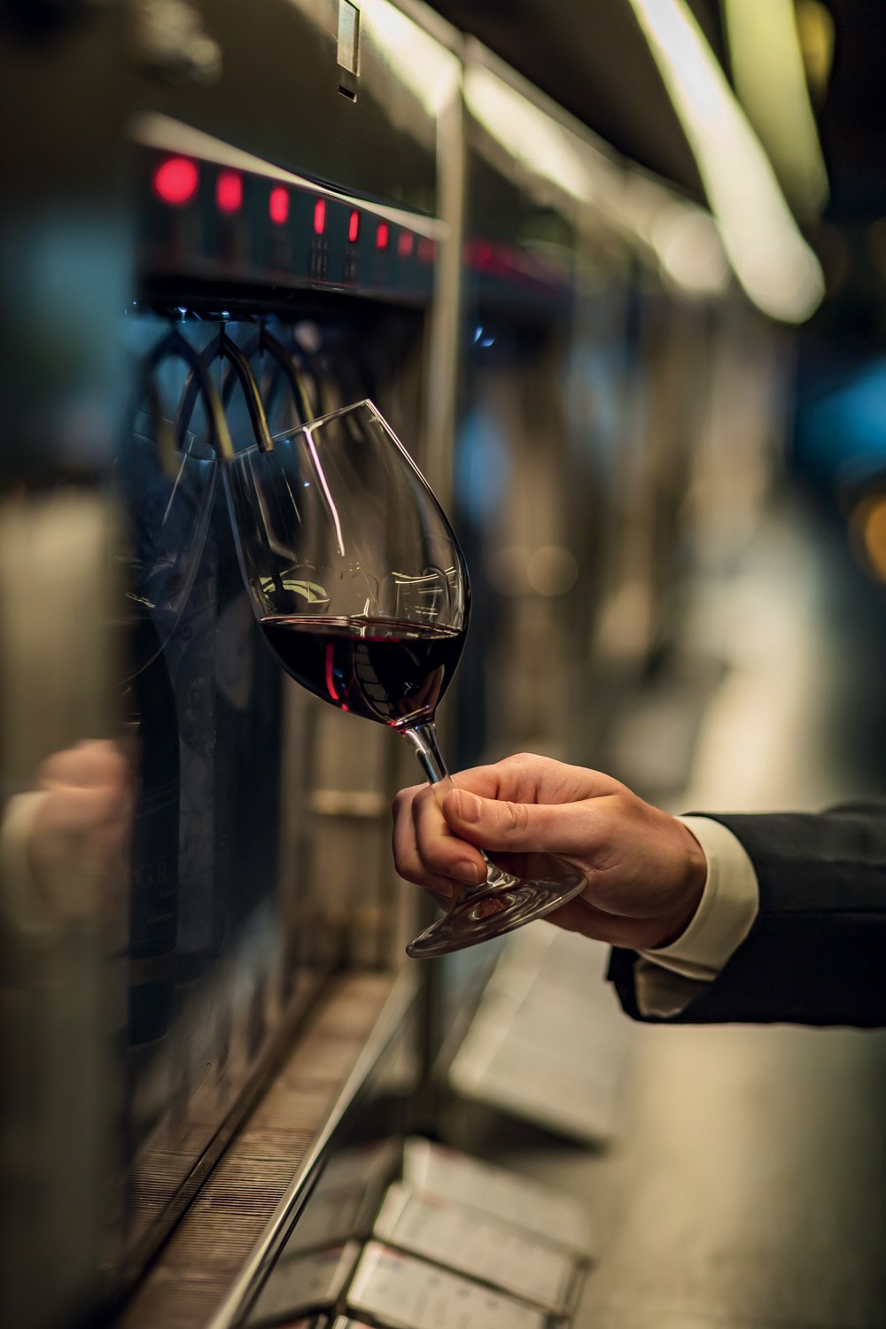 M&atilde;o masculina segurando uma ta&ccedil;a de vinho tinto, sendo servida por uma m&aacute;quina dispensadora com luzes vermelhas em um ambiente escuro