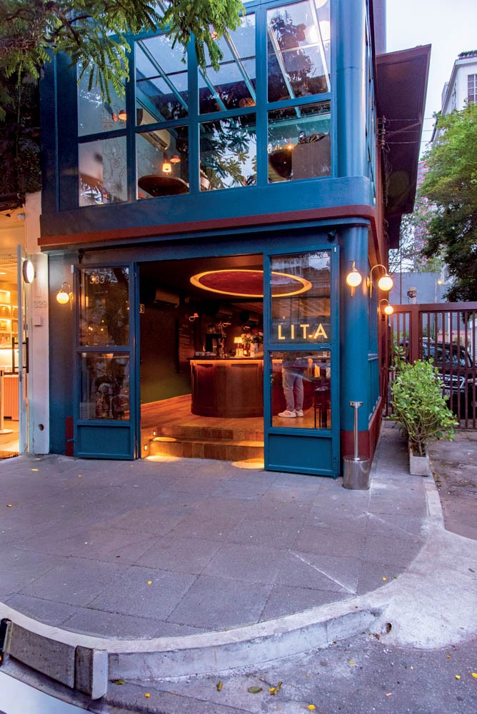Fachada de um bar ou restaurante de dois andares, com paredes azuis escuras e grandes janelas de vidro. O t&eacute;rreo tem portas abertas, revelando um balc&atilde;o e ilumina&ccedil;&atilde;o quente. O andar superior &eacute; todo de vidro, com mesas e cadeiras vis&iacute;veis. Na cal&ccedil;ada, h&aacute; um pequeno arbusto e um port&atilde;o de metal. O letreiro na porta do t&eacute;rreo exibe LIT.A.