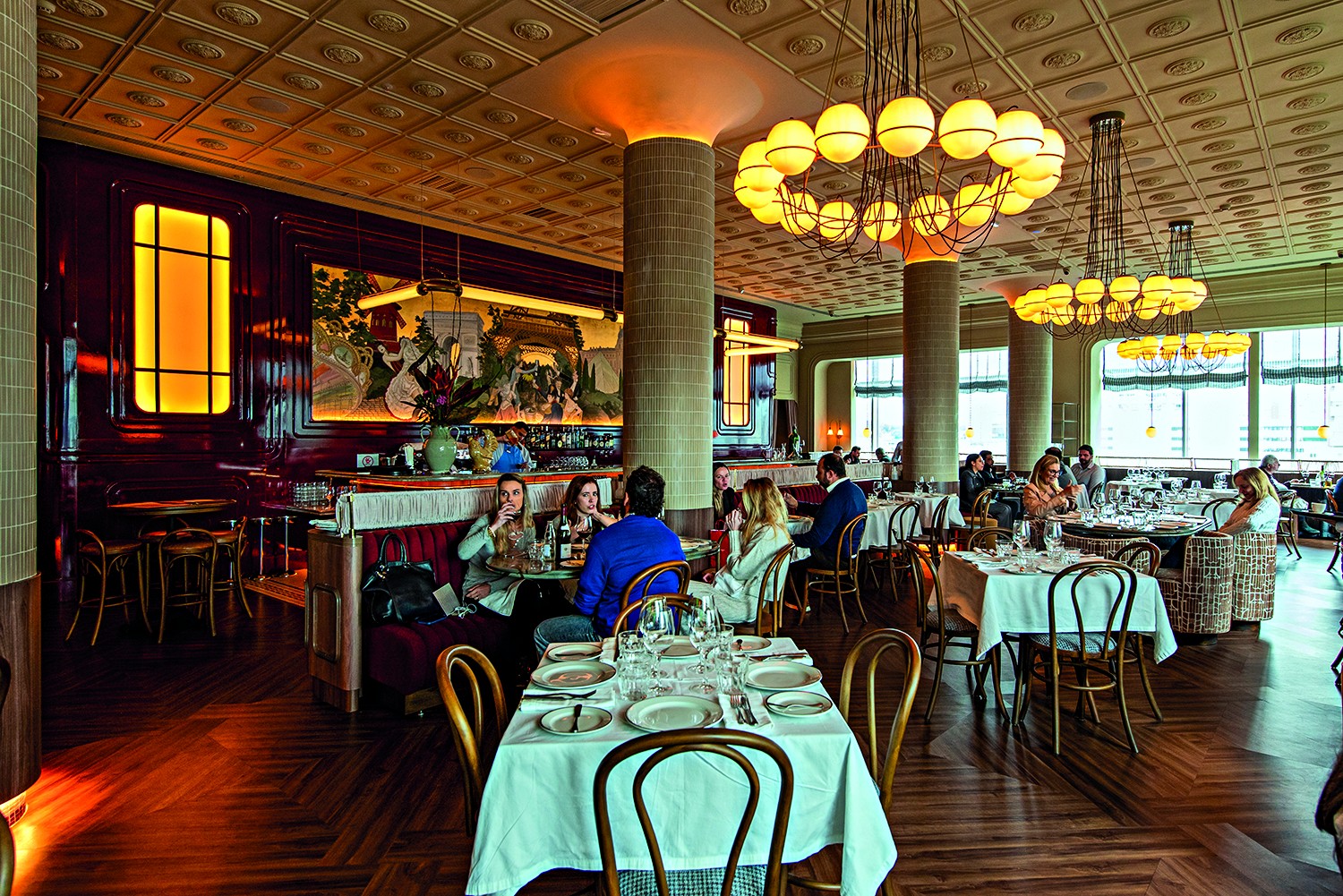 Interior de um restaurante sofisticado com teto dourado e lustres circulares de esferas iluminadas. Pessoas sentadas em mesas com toalhas brancas e cadeiras de madeira, conversando e comendo. Ao fundo, um balc&atilde;o com um grande mural e um barman. O ambiente &eacute; elegante e acolhedor, com ilumina&ccedil;&atilde;o quente e piso de madeira.