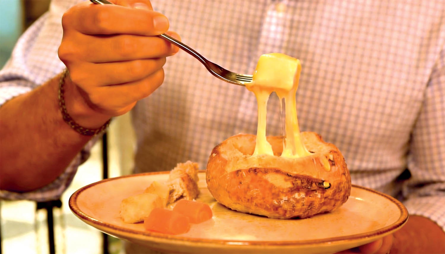 Pessoa com camisa xadrez segurando um garfo com um peda&ccedil;o de p&atilde;o mergulhado em fondue de queijo, que escorre do p&atilde;o para dentro de um p&atilde;o italiano oco, servido em um prato com outros peda&ccedil;os de p&atilde;o e frutas cristalizadas