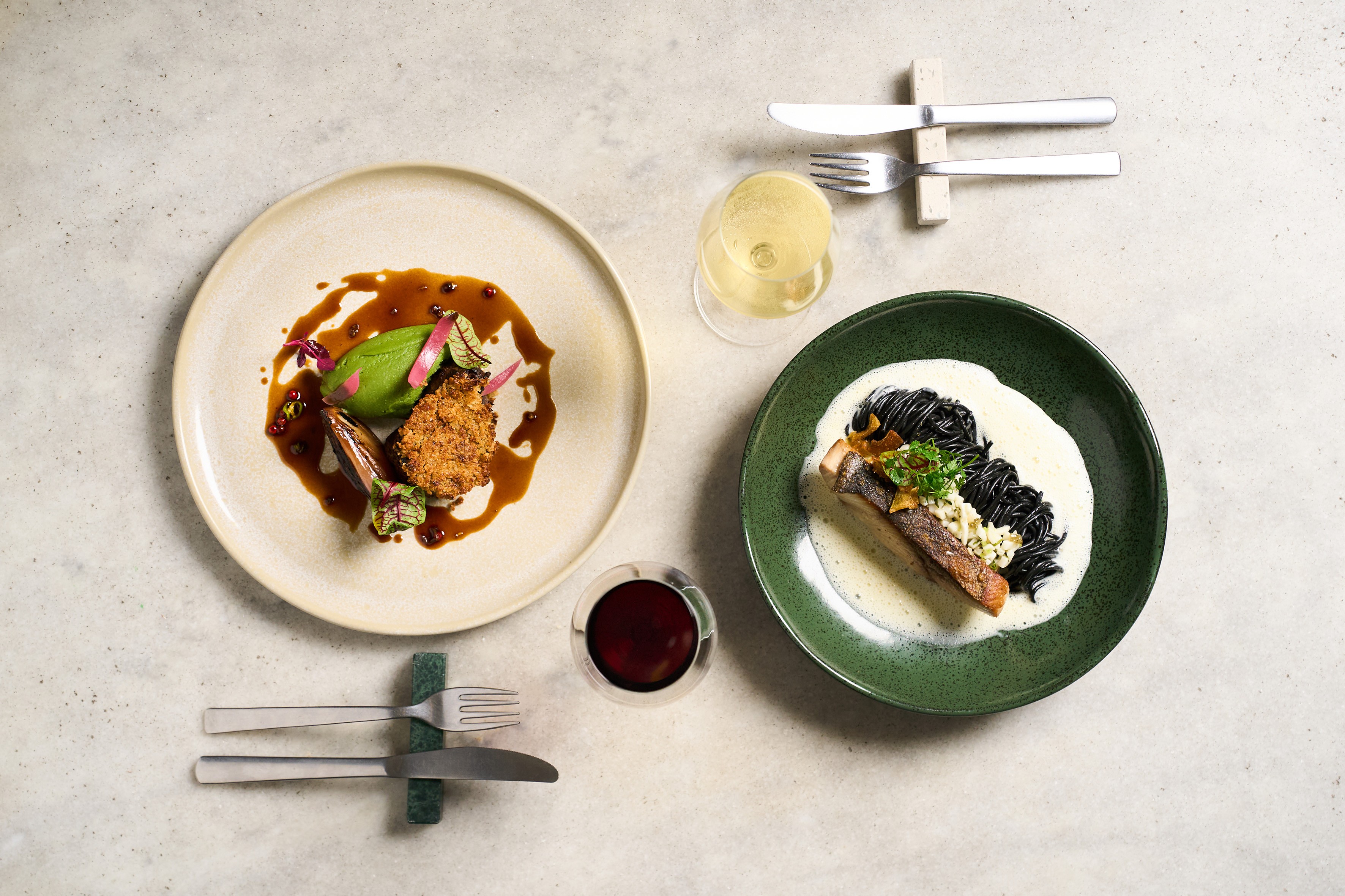 Dois pratos gourmet em uma mesa clara. &Agrave; esquerda, um prato bege com carne empanada, pur&ecirc; verde, molho escuro e folhas. &Agrave; direita, um prato verde com peixe, massa preta, molho branco e temperos. H&aacute; ta&ccedil;as de vinho branco e tinto, e talheres.