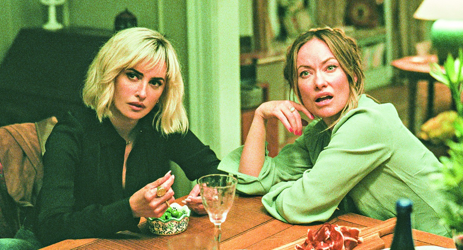 Duas mulheres em uma mesa de madeira. A loira &agrave; esquerda, de blusa preta, segura um petisco verde. A morena &agrave; direita, de blusa verde, tem express&atilde;o surpresa e boca aberta