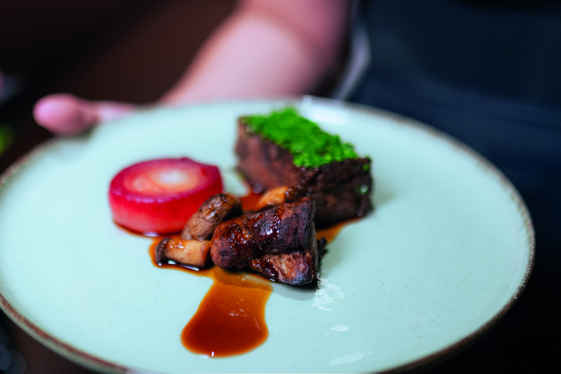 Prato gourmet com carne escura coberta por ervas verdes, cogumelos salteados e uma rodela de beterraba vermelha, servidos em um prato claro com molho marrom