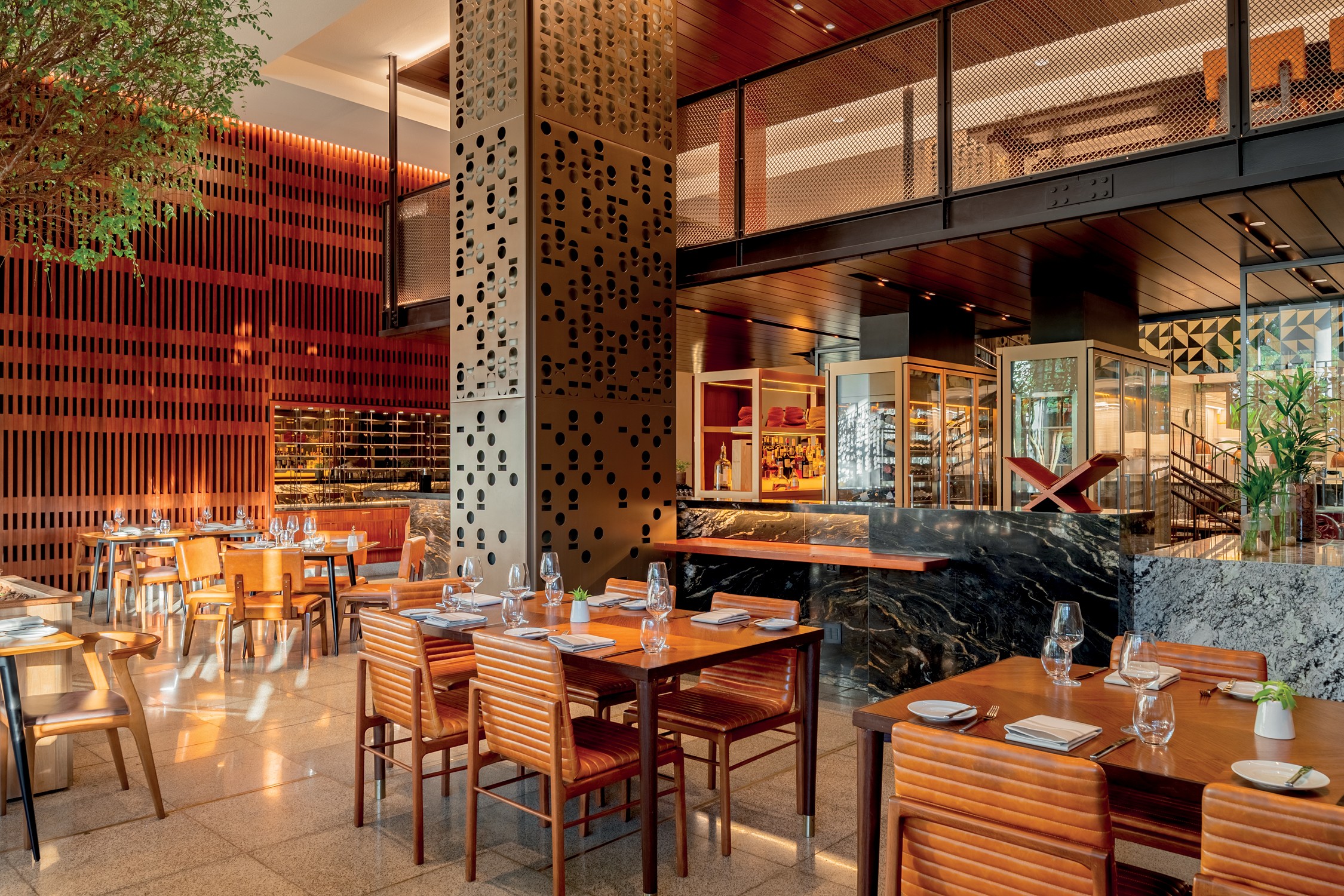 Interior de um restaurante moderno e sofisticado com mesas de madeira e cadeiras estofadas em couro marrom. &Agrave; esquerda, uma parede de madeira ripada e uma &aacute;rvore. &Agrave; direita, um balc&atilde;o de m&aacute;rmore escuro com garrafas de bebidas e uma &aacute;rea de mezanino com grade de metal. O ambiente &eacute; iluminado e elegante