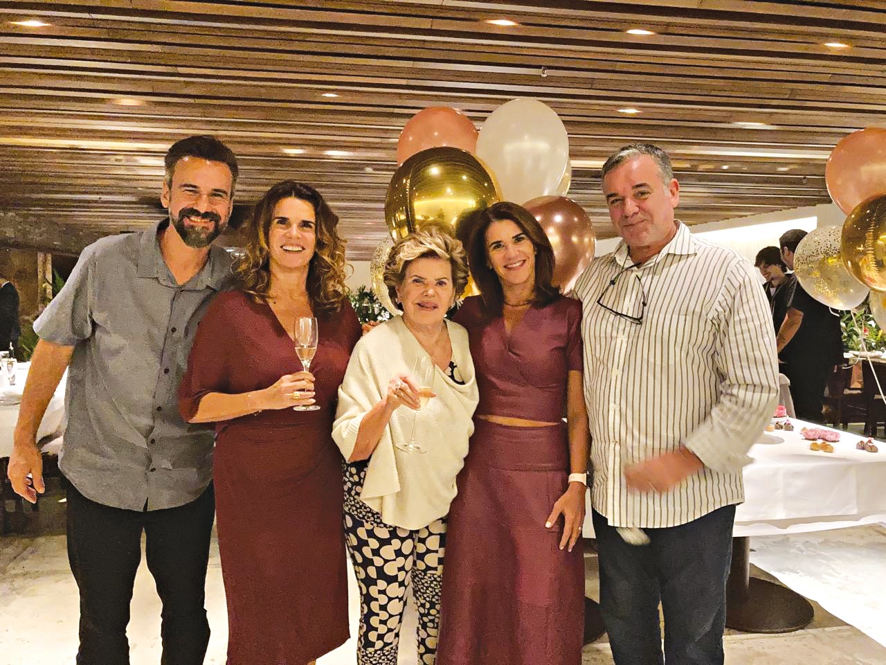 Cinco pessoas sorrindo para a c&acirc;mera em um evento, com bal&otilde;es dourados e ros&ecirc; ao fundo. Um homem de barba e camisa cinza, duas mulheres de vestido vinho, uma senhora de blusa creme e cal&ccedil;a estampada, e um homem de camisa listrada e &oacute;culos pendurados