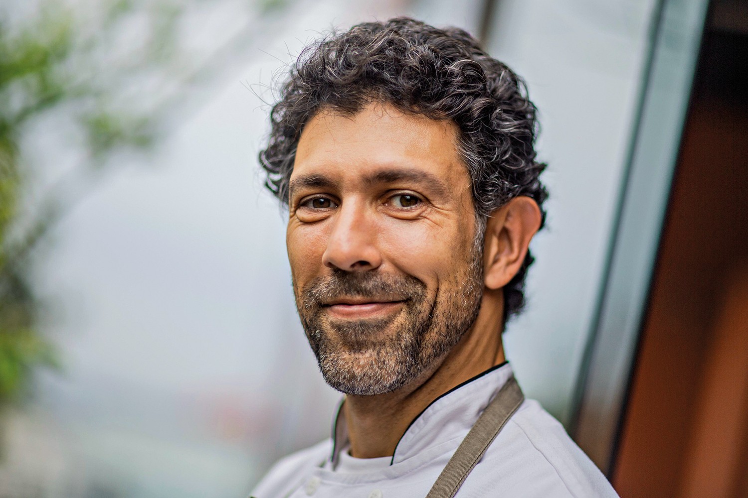 Homem de pele morena, cabelo encaracolado escuro e barba grisalha, sorri levemente para a c&acirc;mera. Ele veste um d&oacute;lm&atilde; branco de chef e um avental marrom, com um fundo desfocado de tons claros e verdes