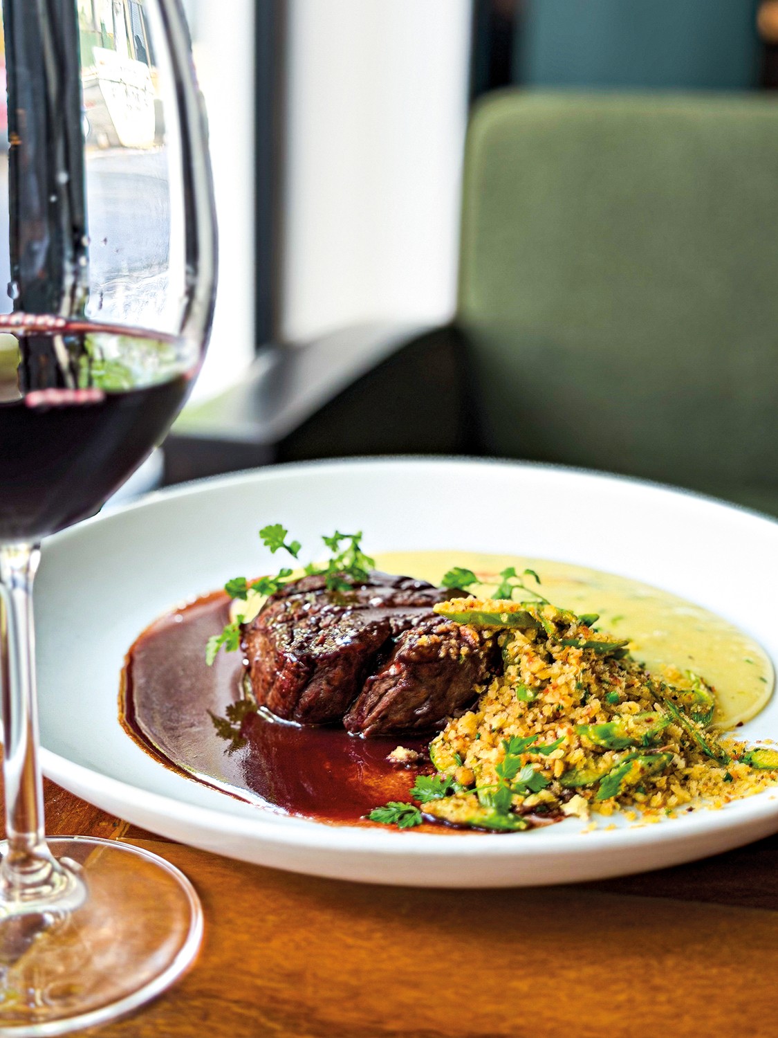 Prato gourmet com medalh&otilde;es de carne ao molho escuro, pur&ecirc; amarelo e farofa verde com ervas frescas, ao lado de uma ta&ccedil;a de vinho tinto, sobre uma mesa de madeira