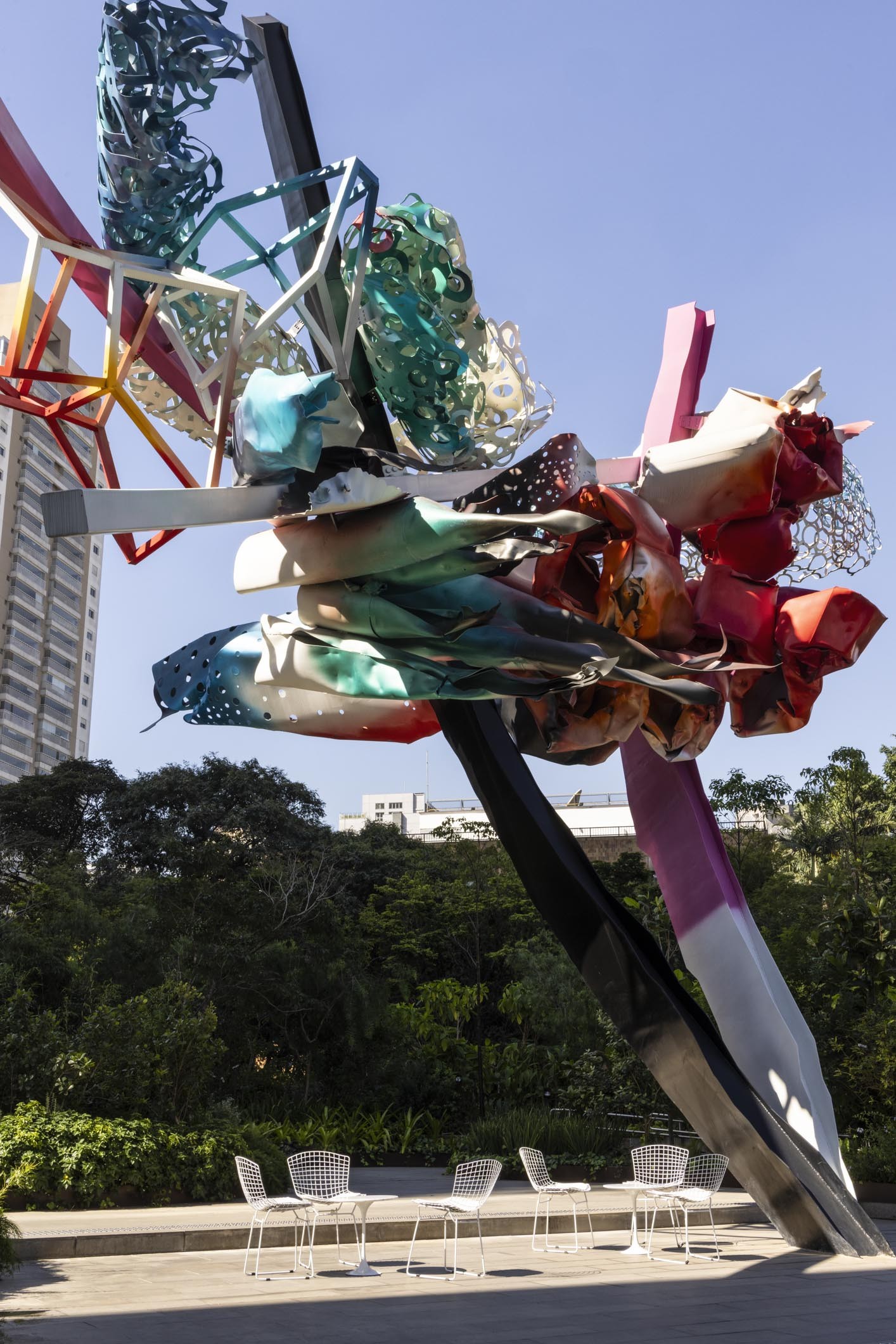 Escultura abstrata colorida, com formas geom&eacute;tricas e org&acirc;nicas em tons de azul, verde, vermelho e branco, sob c&eacute;u azul. Abaixo, quatro mesas brancas com cadeiras de metal vazadas, em um p&aacute;tio de concreto. Ao fundo, &aacute;rvores e um pr&eacute;dio alto.