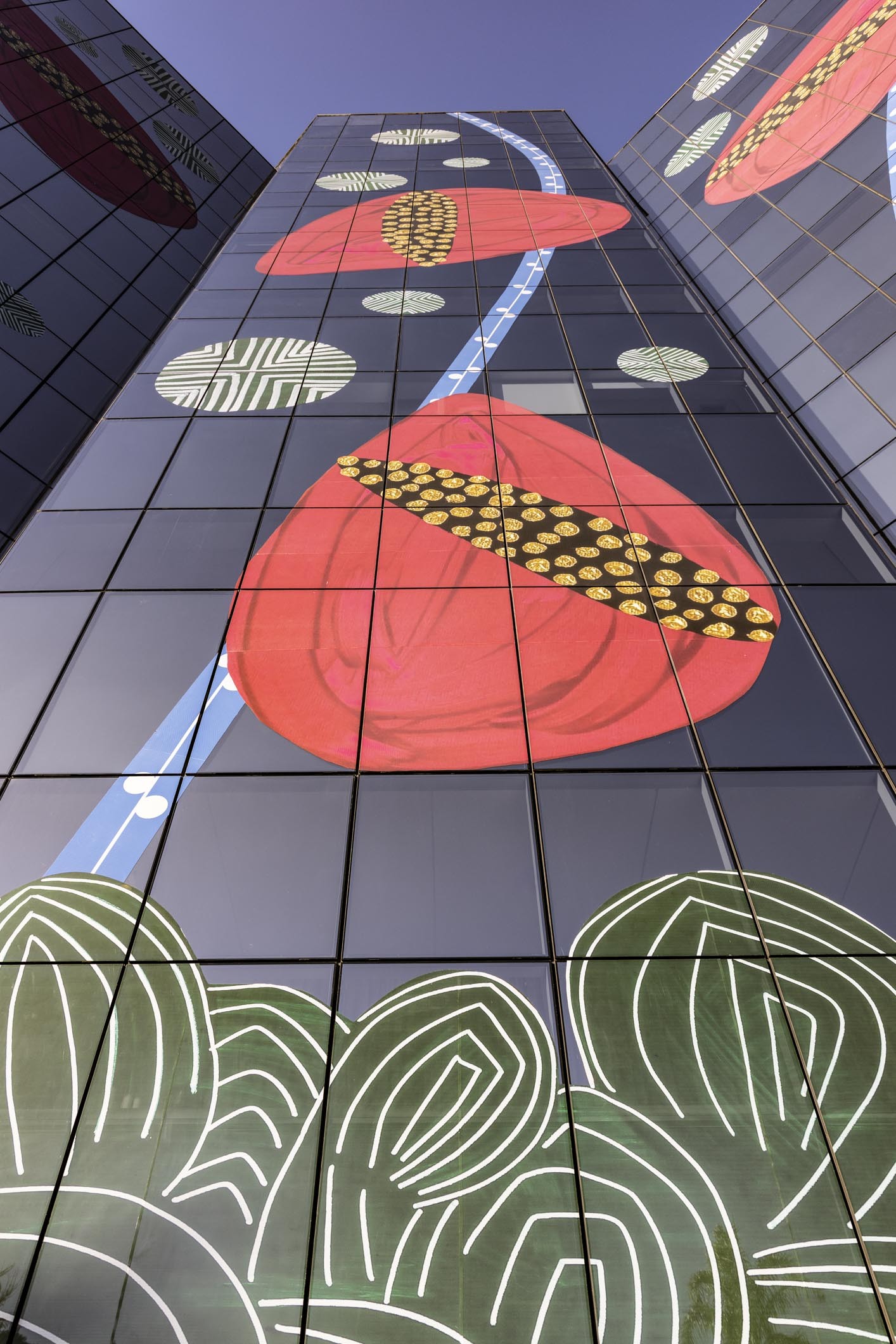Fachada de pr&eacute;dio com janelas espelhadas refletindo um c&eacute;u azul e decorada com arte abstrata. Grandes formas vermelhas com faixas pretas e pontos dourados, linhas azuis diagonais com c&iacute;rculos brancos, e formas verdes com contornos brancos se destacam.