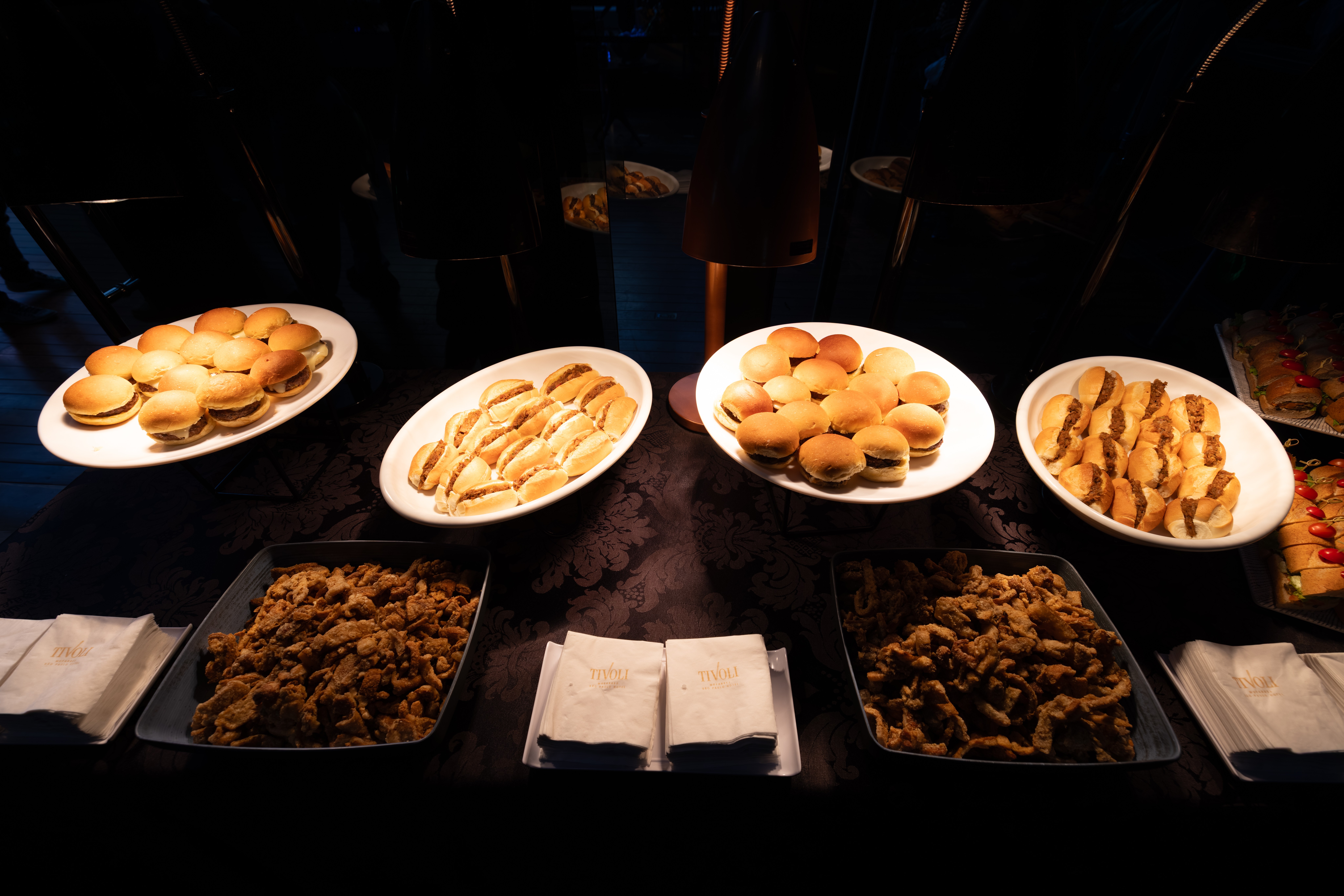 Mesa de buffet com quatro travessas brancas cheias de mini sandu&iacute;ches e duas travessas retangulares escuras com salgados fritos, al&eacute;m de guardanapos de papel dobrados