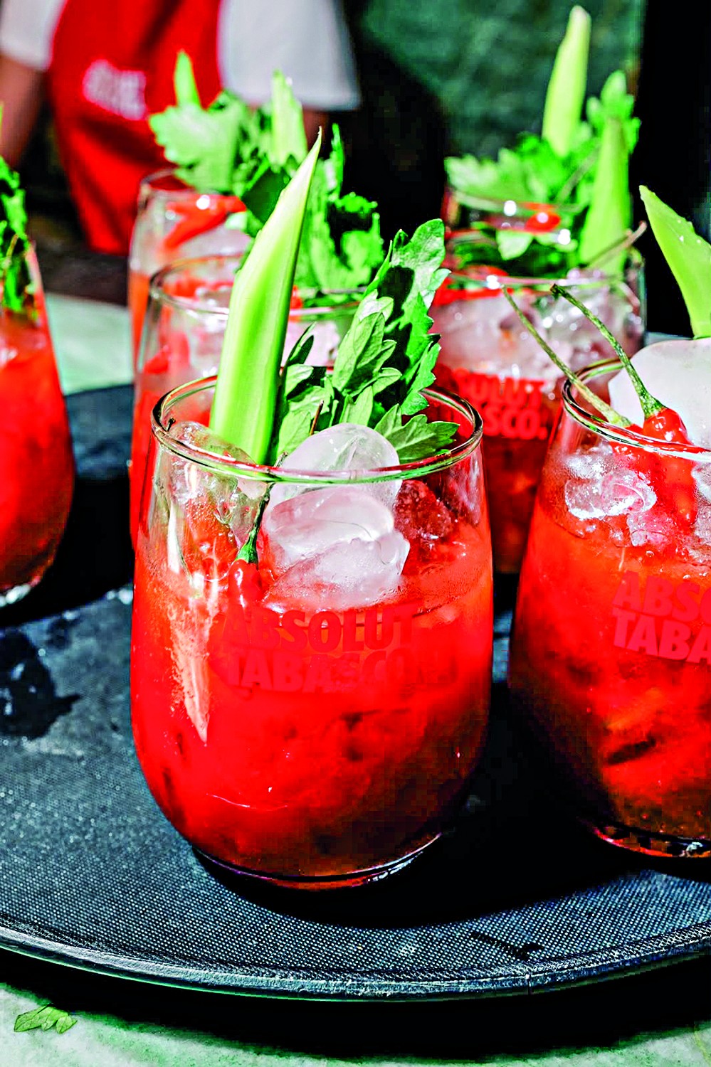 V&aacute;rios copos de Bloody Mary em uma bandeja escura, com gelo, talos de aipo e folhas verdes, alguns com pimenta vermelha. O l&iacute;quido vermelho vibrante preenche os copos, e o nome ABSOLUT TABASCO &eacute; vis&iacute;vel em alguns deles