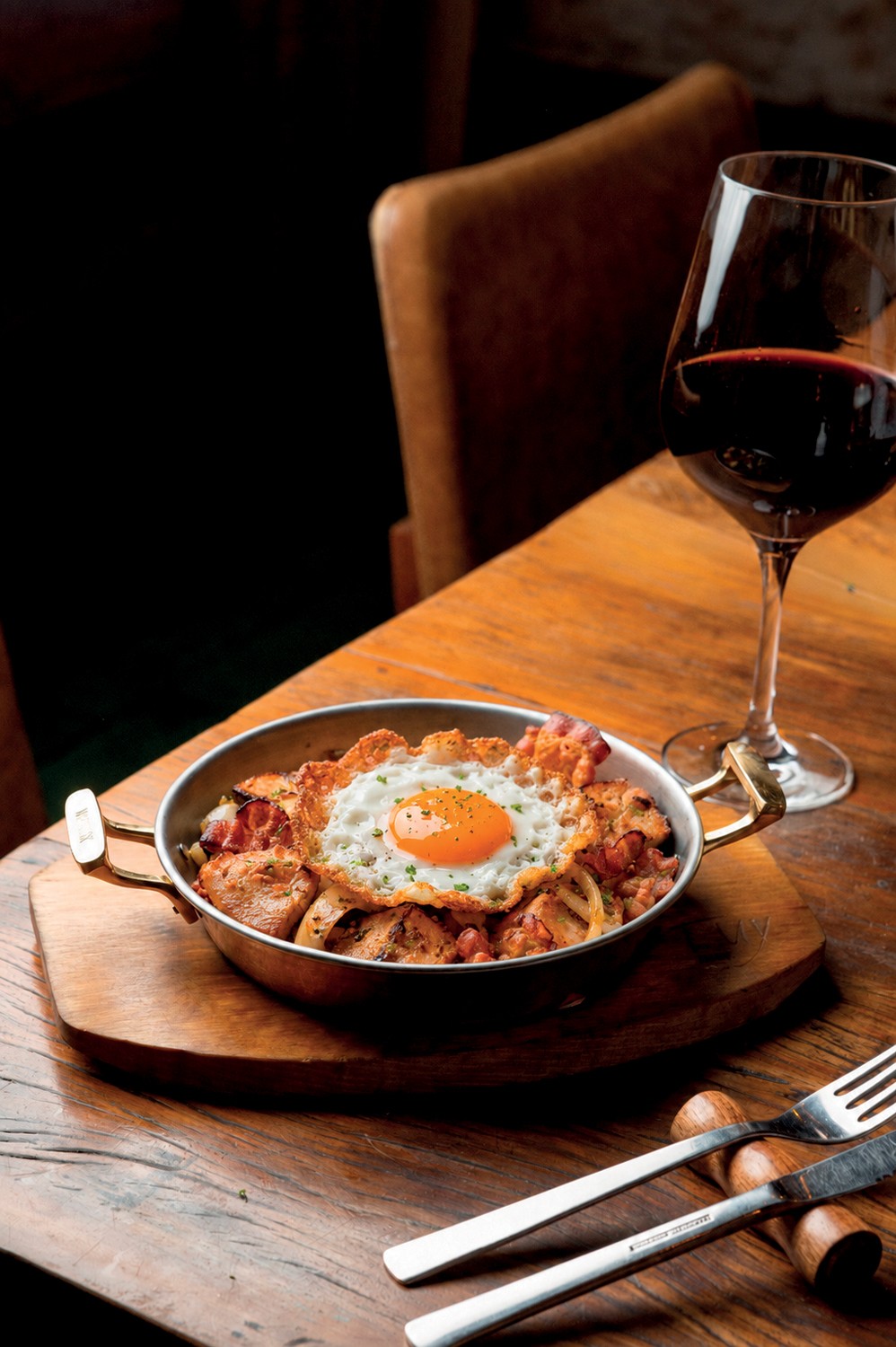 Uma frigideira de metal com al&ccedil;as douradas, contendo um prato com carne, bacon crocante e um ovo frito com gema mole, servido sobre uma t&aacute;bua de madeira r&uacute;stica. Ao lado, uma ta&ccedil;a de vinho tinto e talheres de metal com cabos de madeira, tudo sobre uma mesa de madeira escura. O fundo &eacute; desfocado e escuro, com um assento de cadeira marrom vis&iacute;vel.