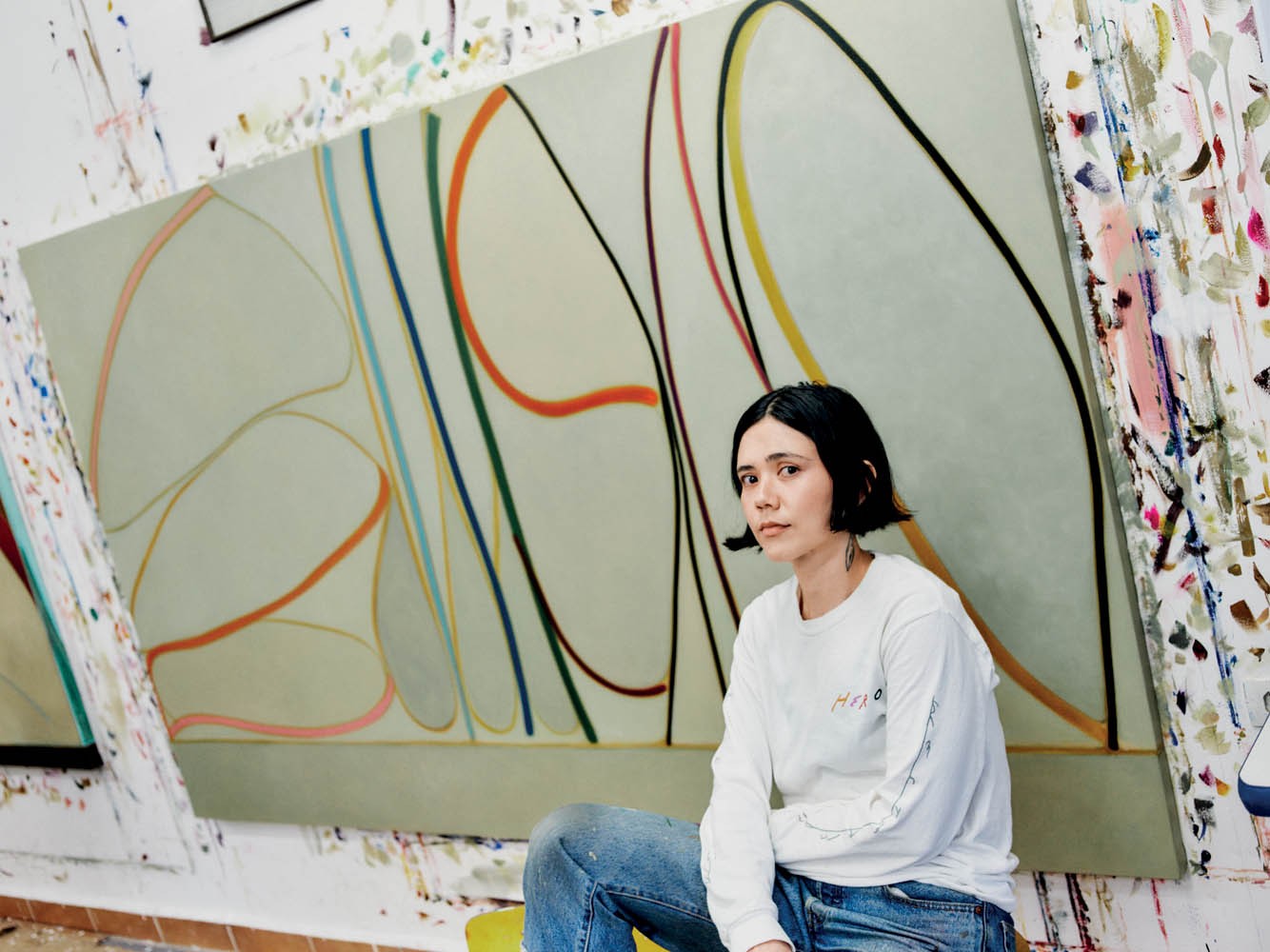 Mulher de cabelo preto curto, blusa branca e jeans, sentada em um ateli&ecirc; com paredes manchadas de tinta, ao lado de uma grande tela abstrata com linhas coloridas e formas org&acirc;nicas