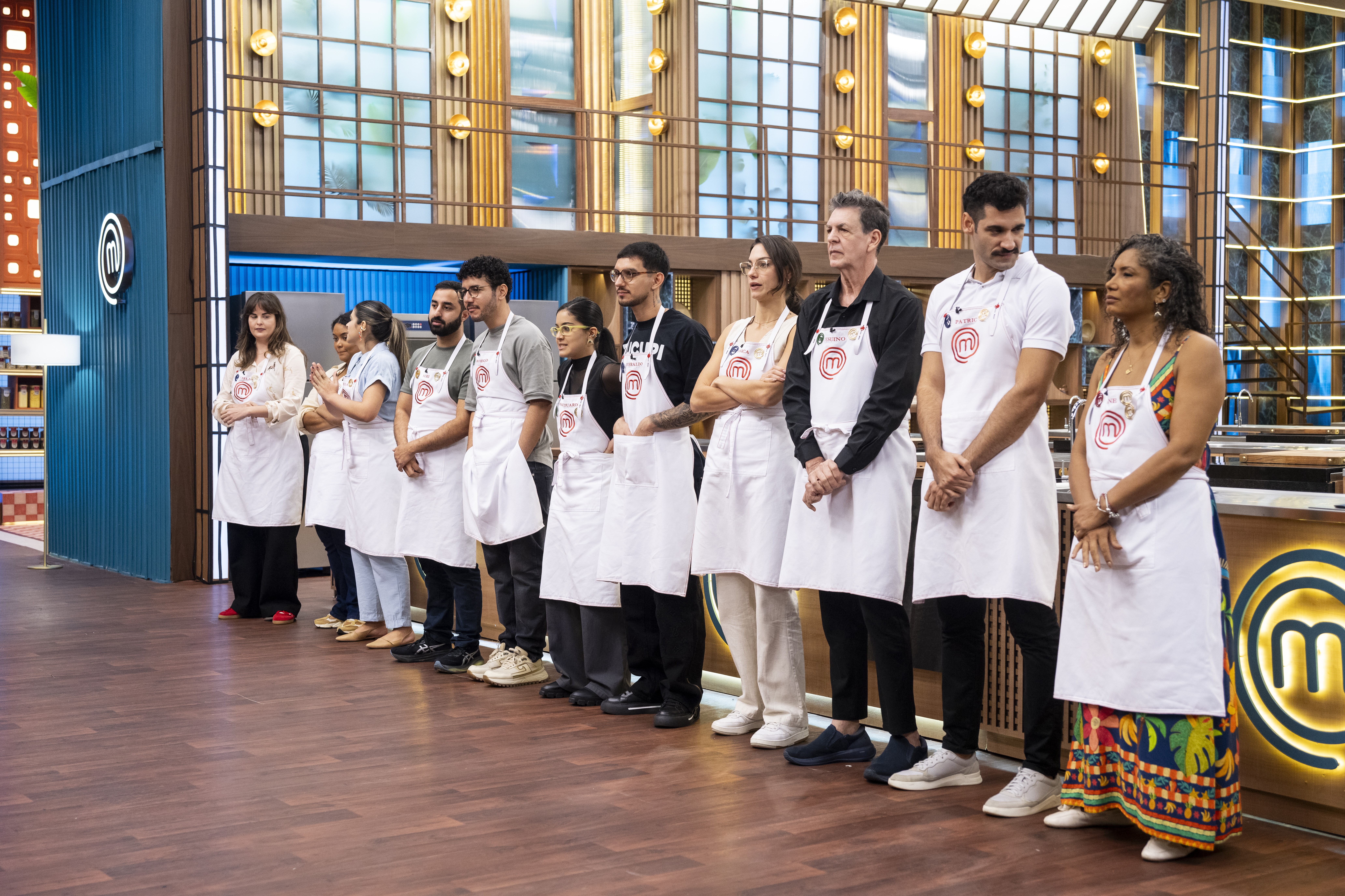 Um grupo de dez pessoas, incluindo homens e mulheres de diversas etnias, usando aventais brancos com o logo do MasterChef, est&atilde;o em fila em um est&uacute;dio de televis&atilde;o com paredes de madeira e detalhes em azul. Eles parecem atentos e s&eacute;rios, aguardando instru&ccedil;&otilde;es.