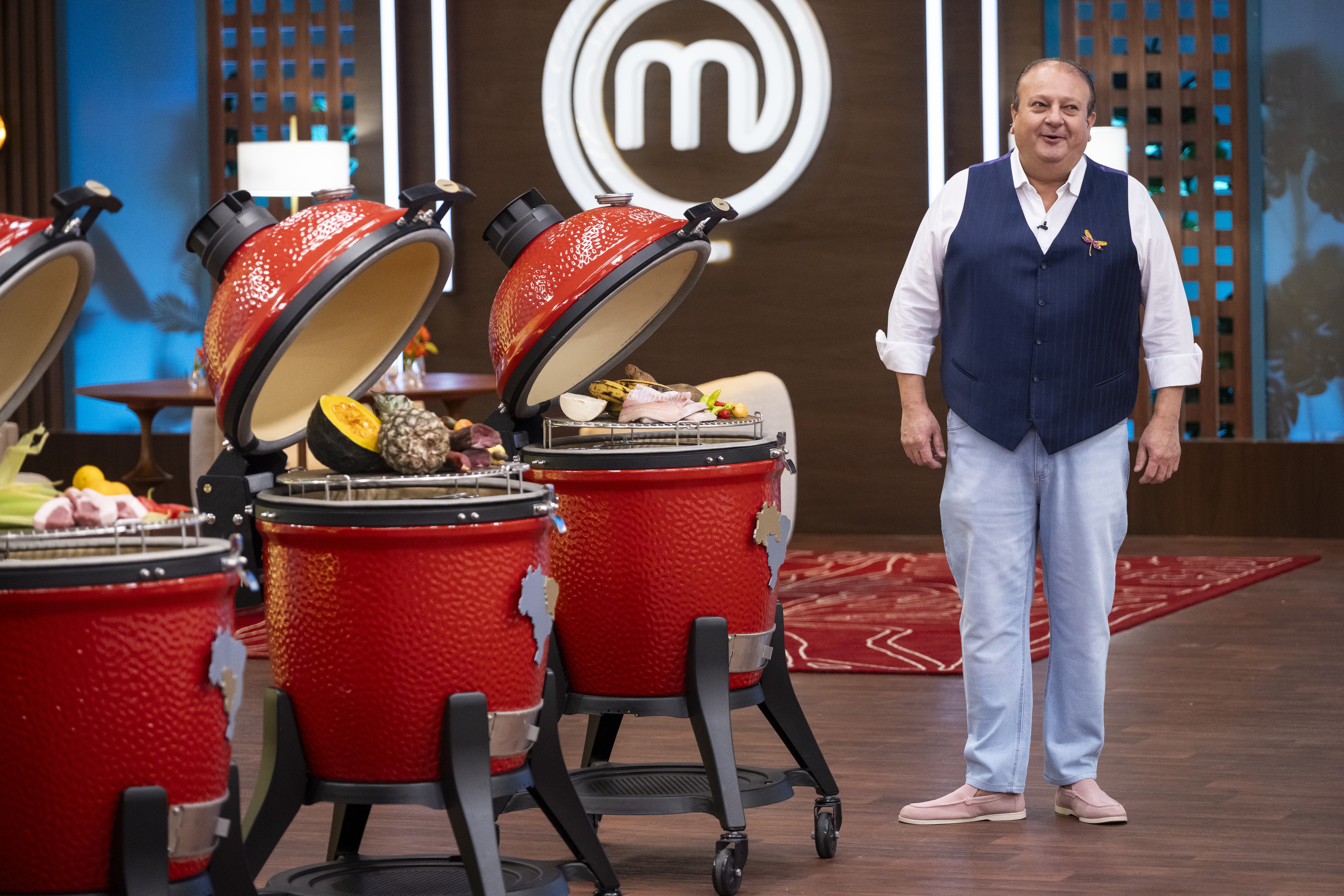 Homem sorridente de camisa branca, colete azul e cal&ccedil;a clara, em p&eacute; ao lado de tr&ecirc;s churrasqueiras vermelhas com alimentos crus, no cen&aacute;rio do MasterChef