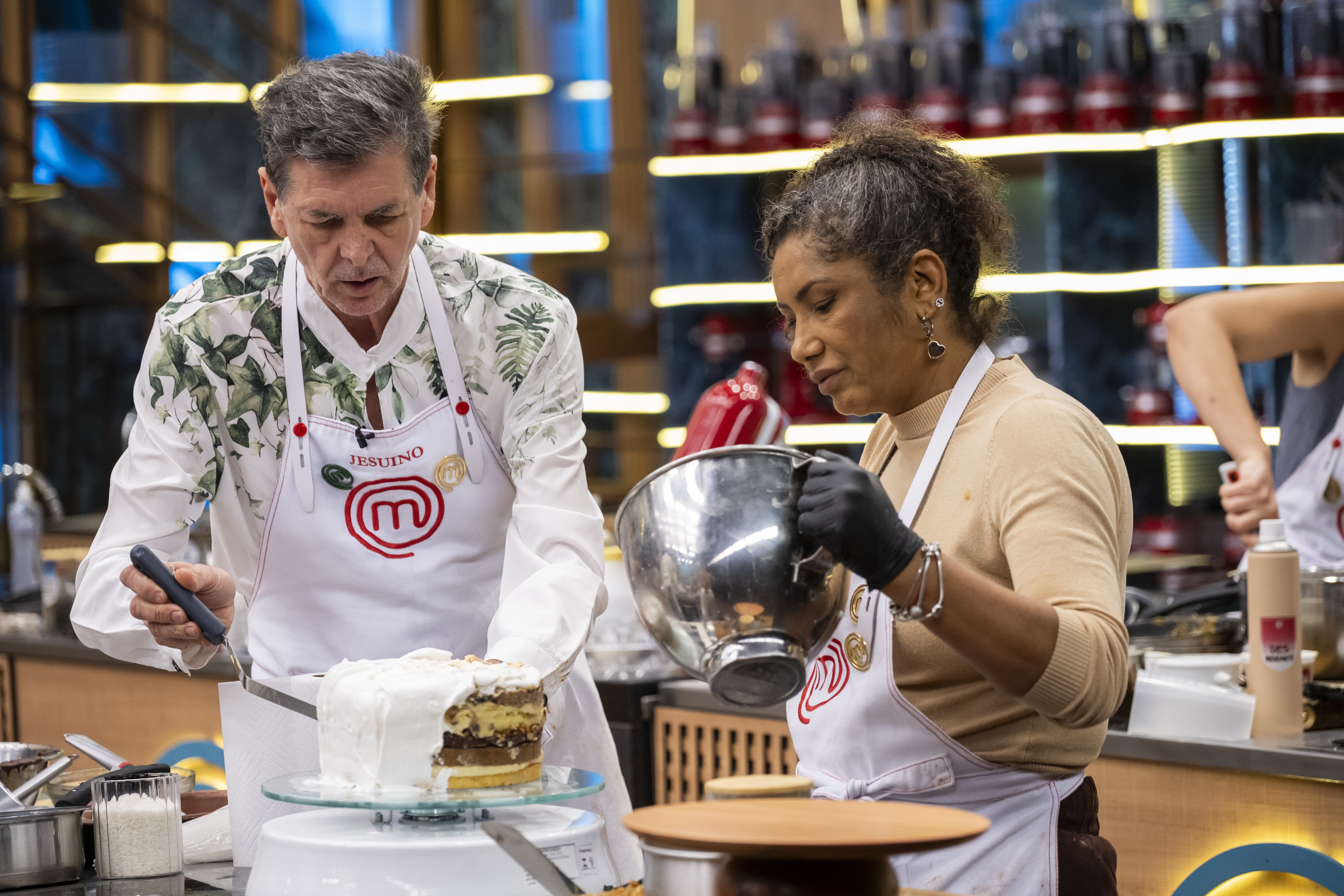 Um homem e uma mulher em aventais brancos de chef preparam um bolo. O homem, &agrave; esquerda, usa uma camisa estampada e espalha cobertura branca em um bolo de camadas. A mulher, &agrave; direita, com cabelo cacheado e luvas pretas, segura uma tigela de metal. Ambos est&atilde;o concentrados na tarefa, em uma cozinha profissional com prateleiras iluminadas ao fundo