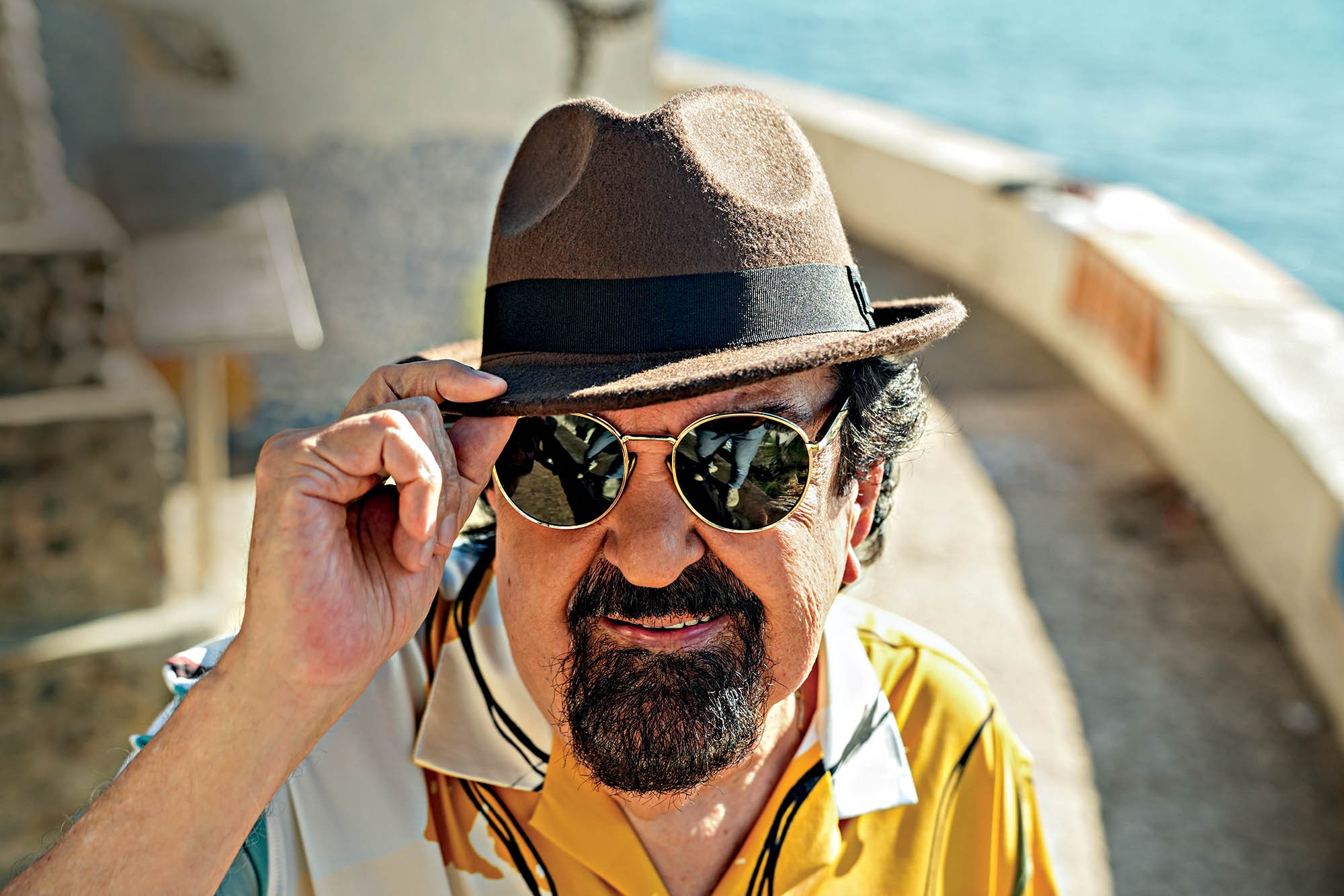 Homem de pele clara, barba e bigode escuros, sorri levemente enquanto segura a aba de seu chap&eacute;u marrom. Ele usa &oacute;culos de sol redondos e uma camisa amarela estampada, com o mar azul ao fundo