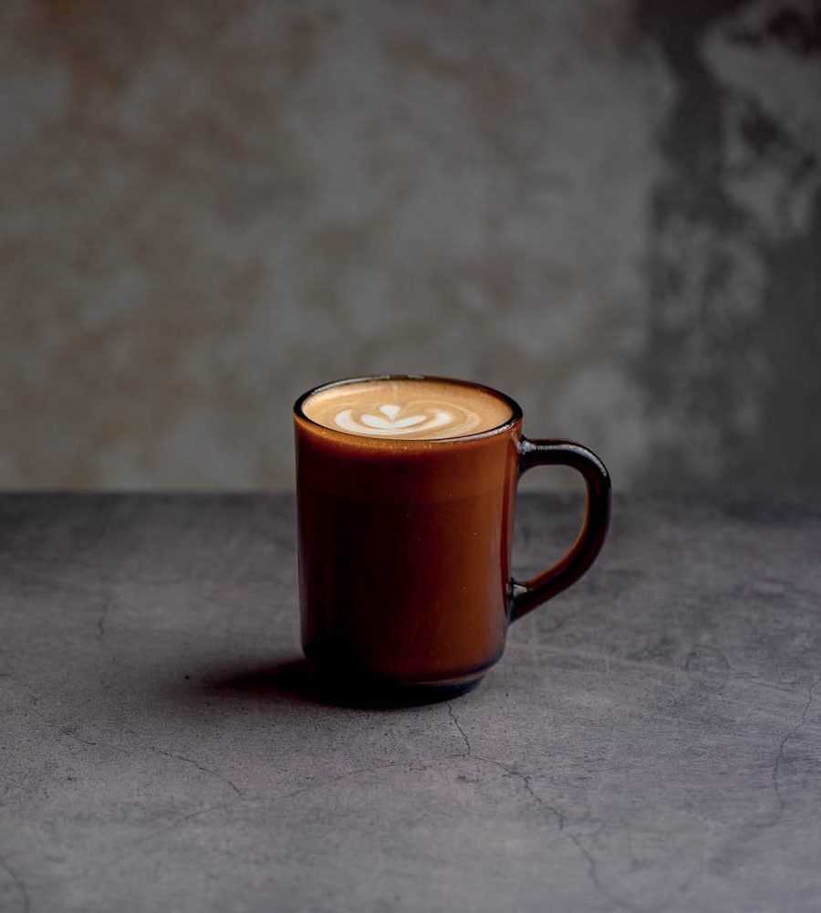 Caneca marrom escura com caf&eacute; latte e arte de cora&ccedil;&atilde;o na espuma branca, sobre uma superf&iacute;cie cinza escura texturizada e fundo bege acinzentado