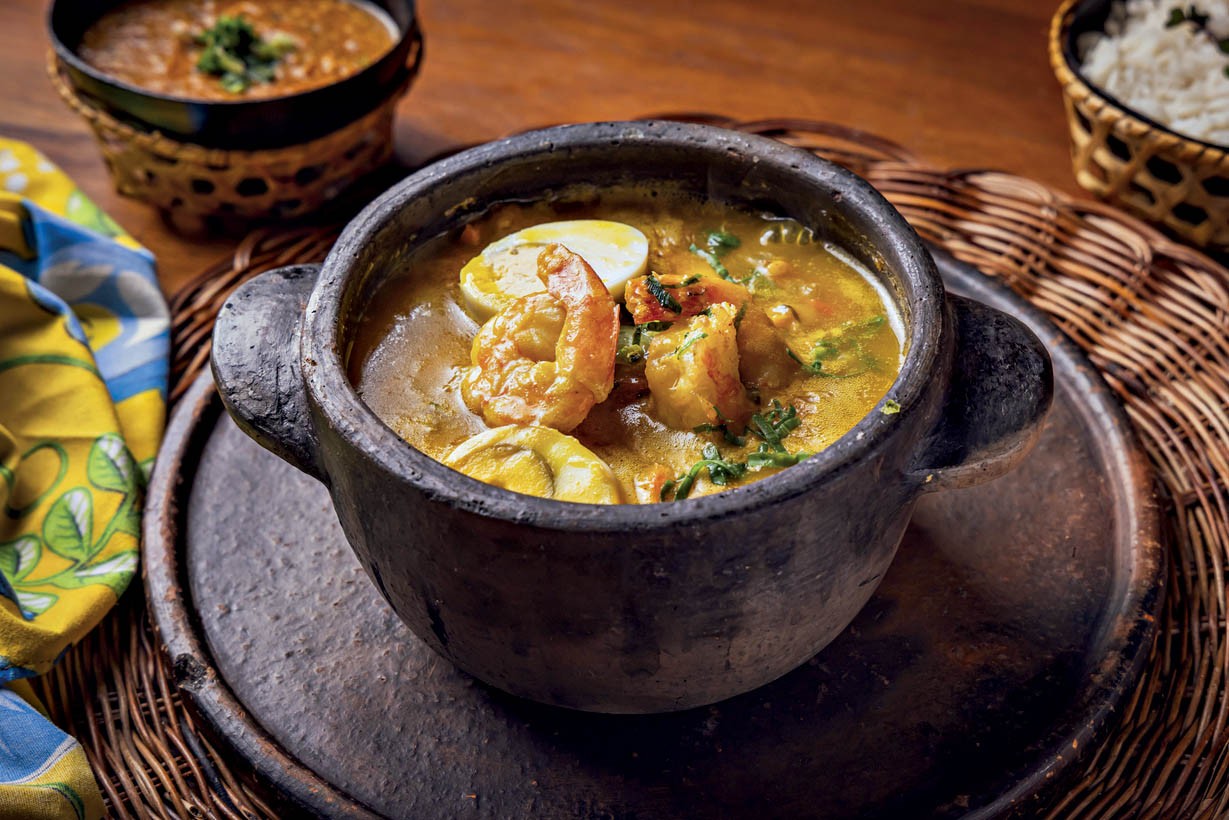 Moqueca de camar&atilde;o com ovos cozidos em panela de barro escura, sobre um prato r&uacute;stico de madeira e esteira de palha. Ao fundo, um pote de arroz branco e outro de pir&atilde;o, ambos em cestos de palha. Um tecido amarelo com detalhes azuis e verdes est&aacute; &agrave; esquerda
