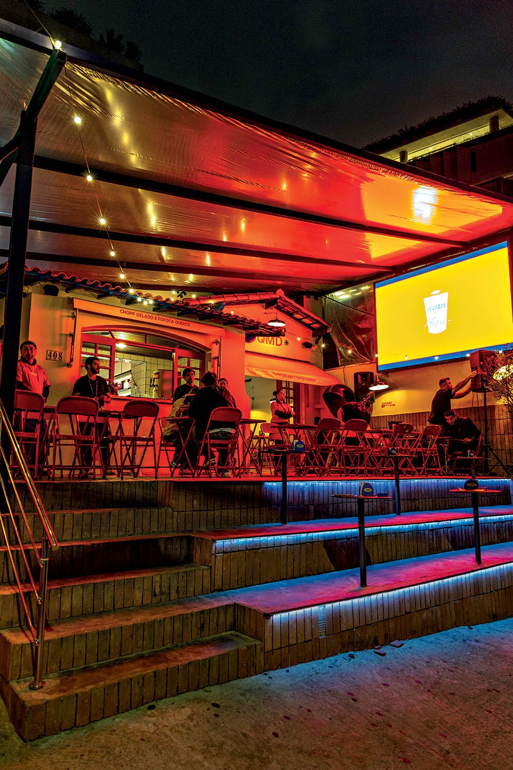 Vista noturna de um bar com teto de lona iluminado por luzes vermelhas e brancas. Clientes sentados em mesas e cadeiras vermelhas, alguns em frente a um balc&atilde;o. Um tel&atilde;o amarelo exibe um copo branco. Escadas com ilumina&ccedil;&atilde;o azul neon levam ao n&iacute;vel superior do bar