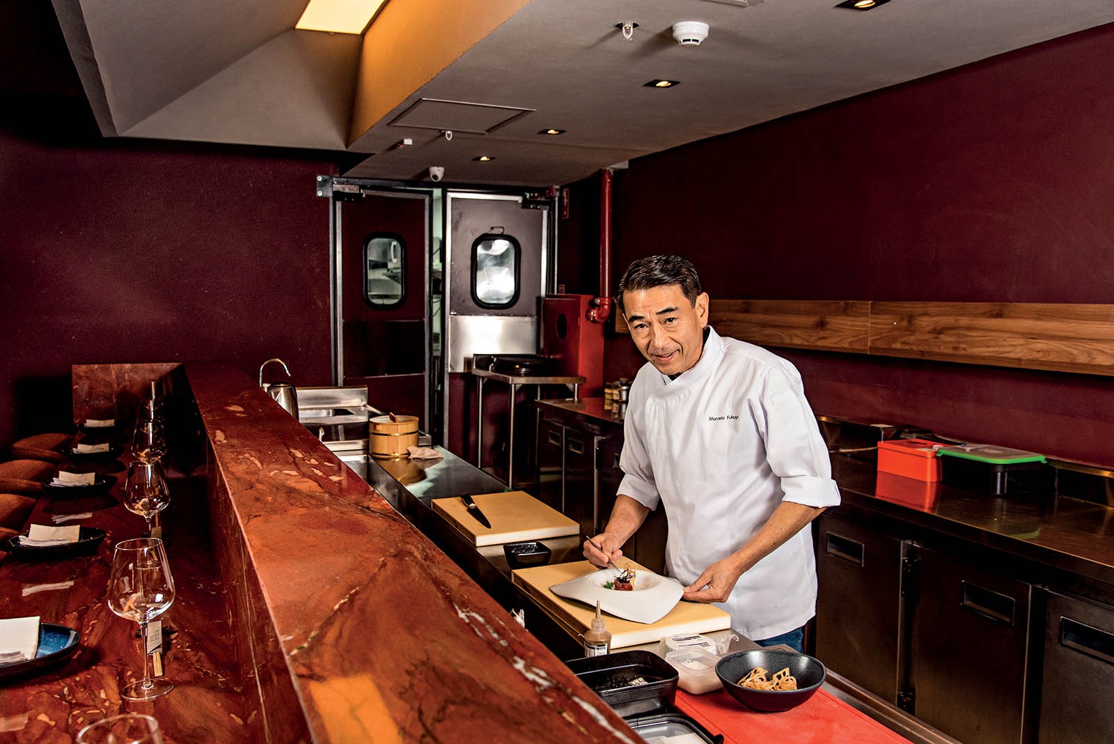 Chef japon&ecirc;s de meia-idade, vestindo d&oacute;lm&atilde; branco, finaliza um prato em balc&atilde;o de cozinha de restaurante com paredes vinho e ilumina&ccedil;&atilde;o quente