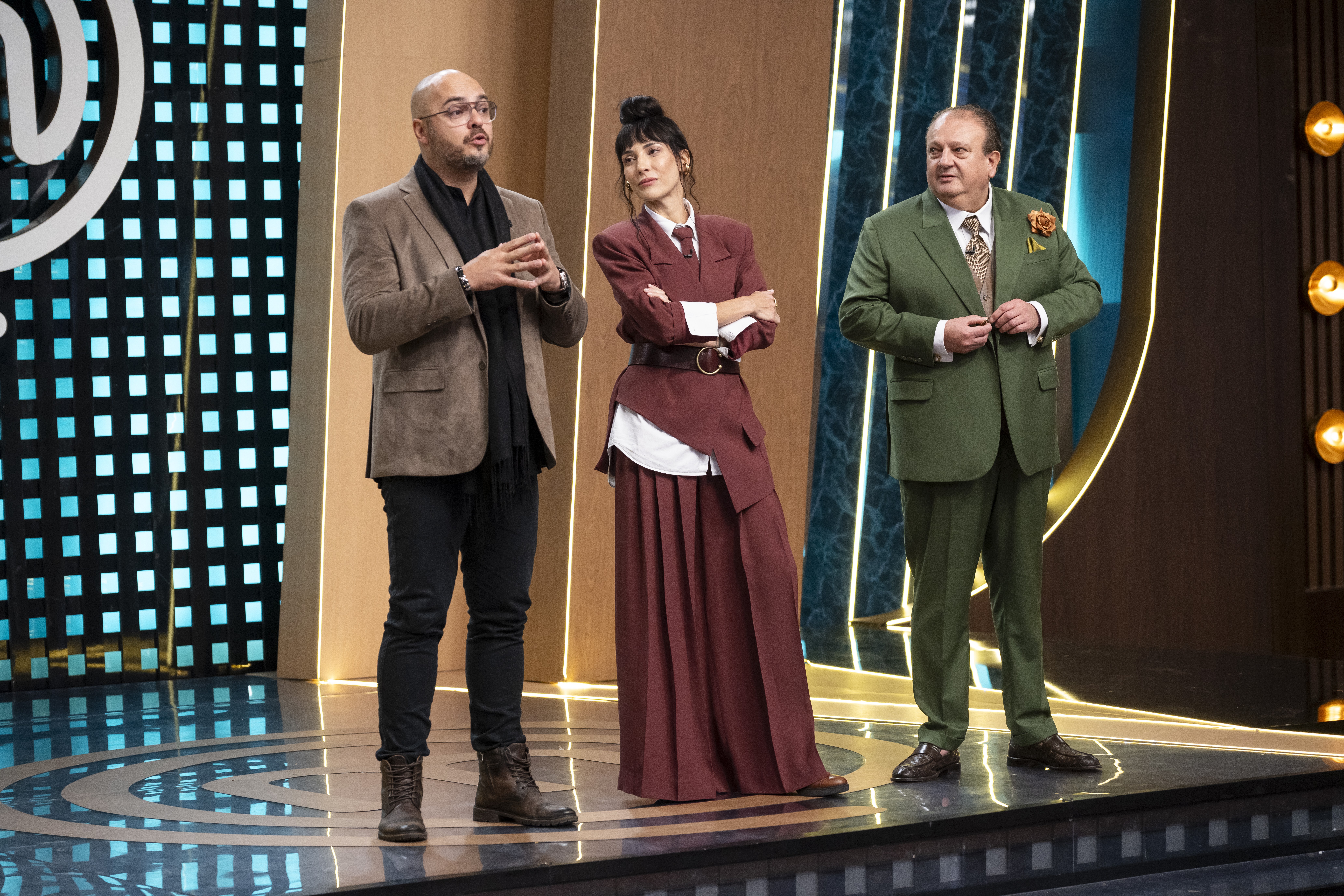 Tr&ecirc;s jurados, dois homens e uma mulher, em um palco de programa de culin&aacute;ria. O homem &agrave; esquerda, careca e de &oacute;culos, veste blazer marrom e cachecol preto, gesticulando. A mulher ao centro usa um conjunto vinho com saia longa e blusa branca, bra&ccedil;os cruzados. O homem &agrave; direita, de terno verde-oliva, abotoa o palet&oacute;. O palco tem ilumina&ccedil;&atilde;o azul e dourada