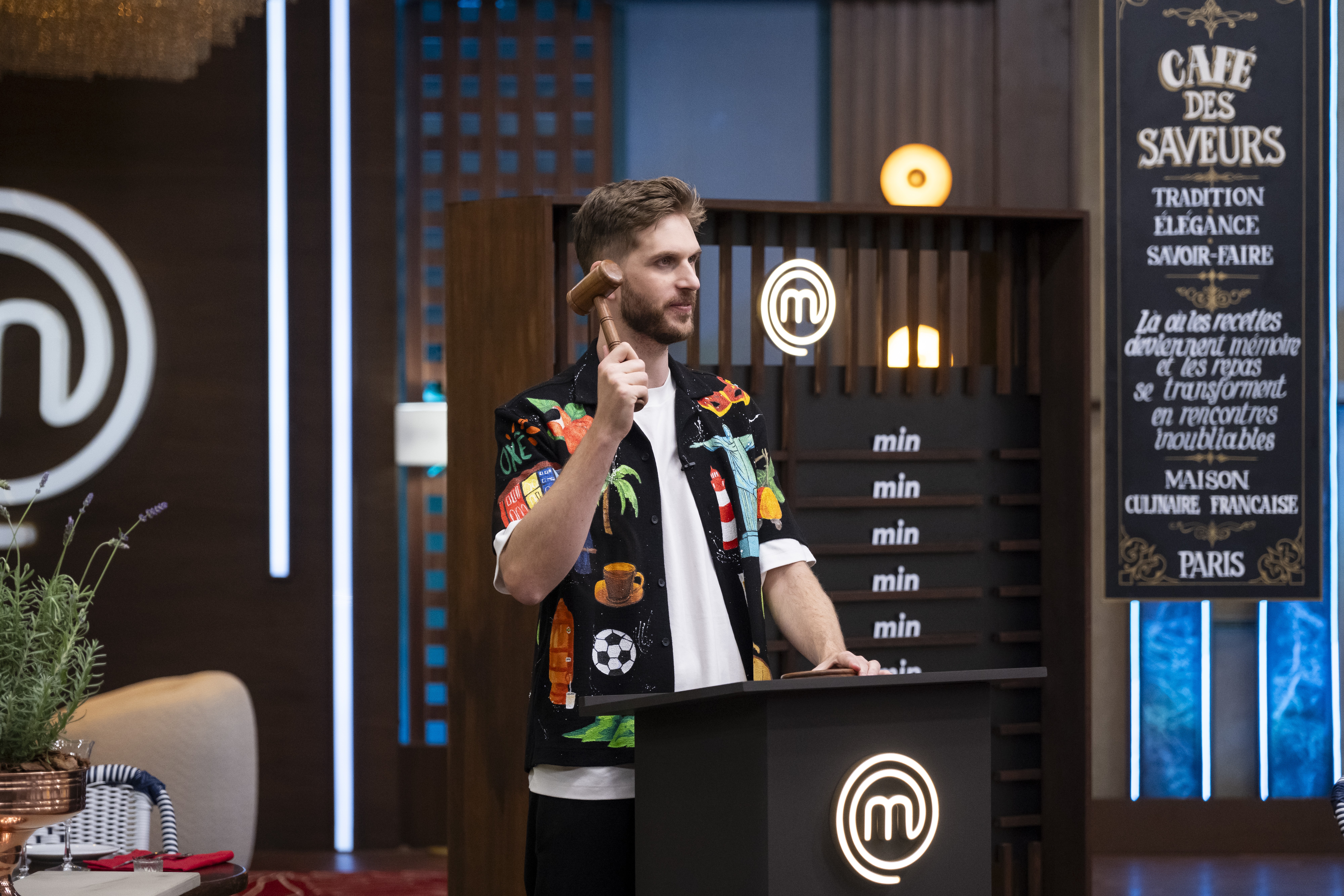 Homem barbudo de camisa estampada colorida e camiseta branca, segurando um martelo de madeira, em p&eacute; atr&aacute;s de um p&uacute;lpito preto com o logo M do MasterChef. Ao fundo, um painel com min repetido e uma placa preta com Caf&eacute; des Saveurs em franc&ecirc;s