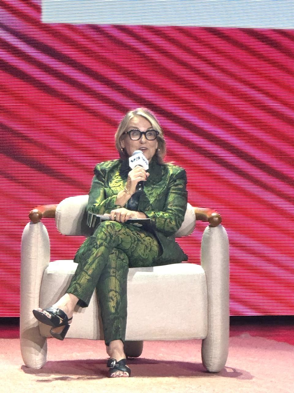 Mulher loira de &oacute;culos, vestindo um conjunto verde estampado, sentada em uma poltrona clara, falando ao microfone em um palco com fundo vermelho listrado