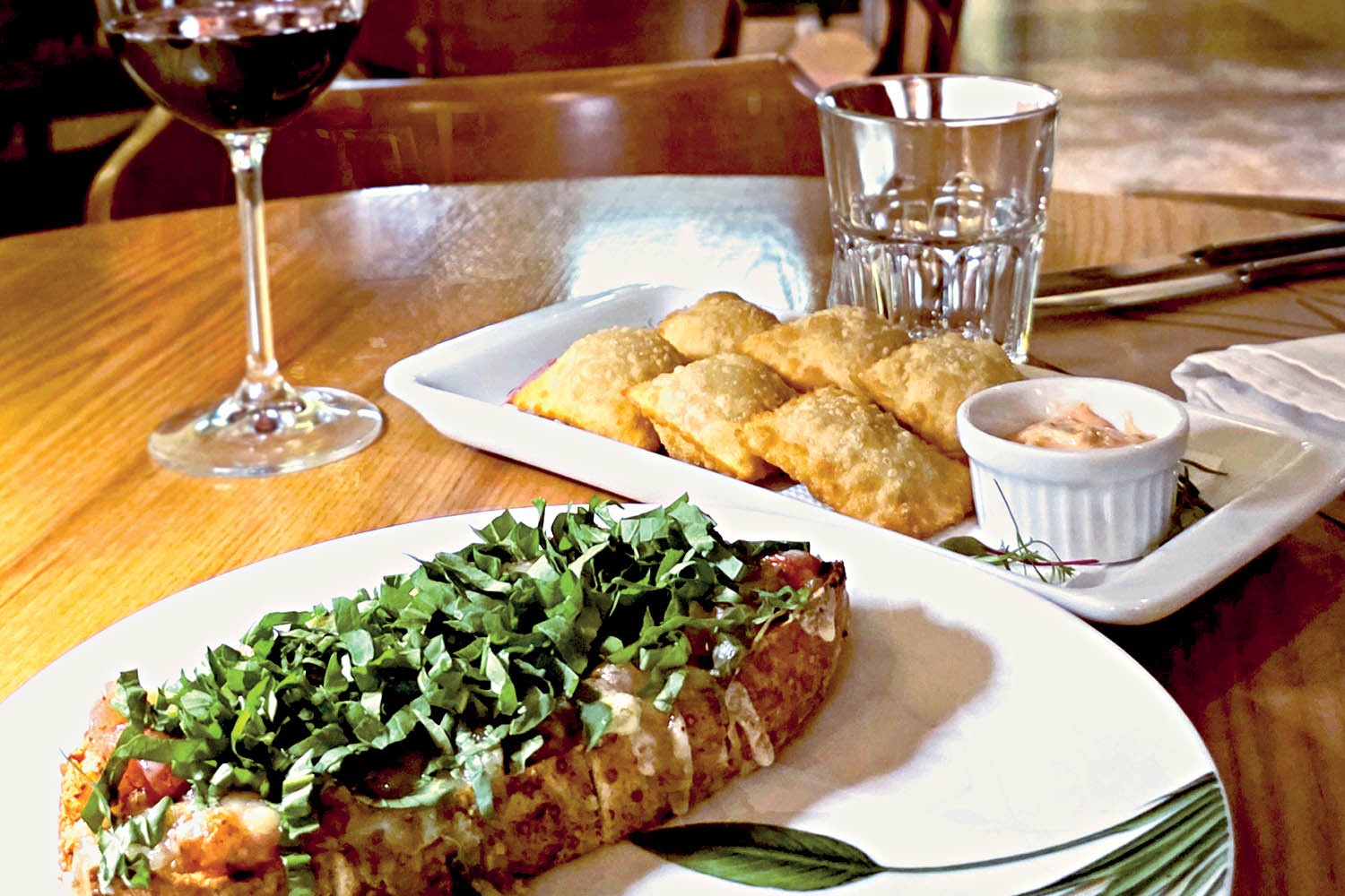 Mesa de madeira com prato branco contendo bruschetta coberta por folhas verdes, e outro prato retangular com past&eacute;is fritos e um molho em ramequim. Ao fundo, uma ta&ccedil;a de vinho tinto e um copo de &aacute;gua.