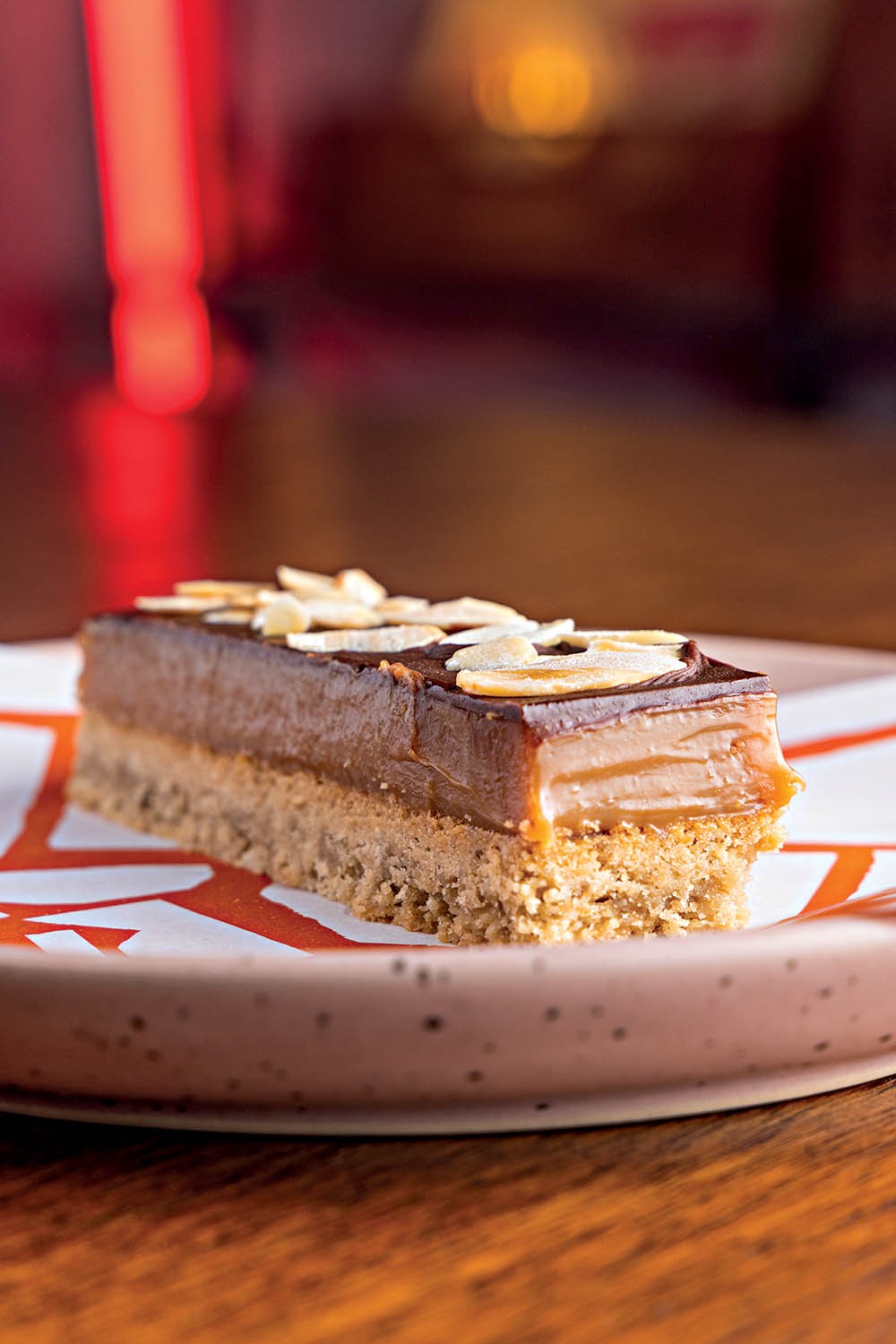 Fatia retangular de torta com base crocante, recheio de doce de leite, cobertura de chocolate e am&ecirc;ndoas laminadas, servida em prato branco com detalhes laranja
