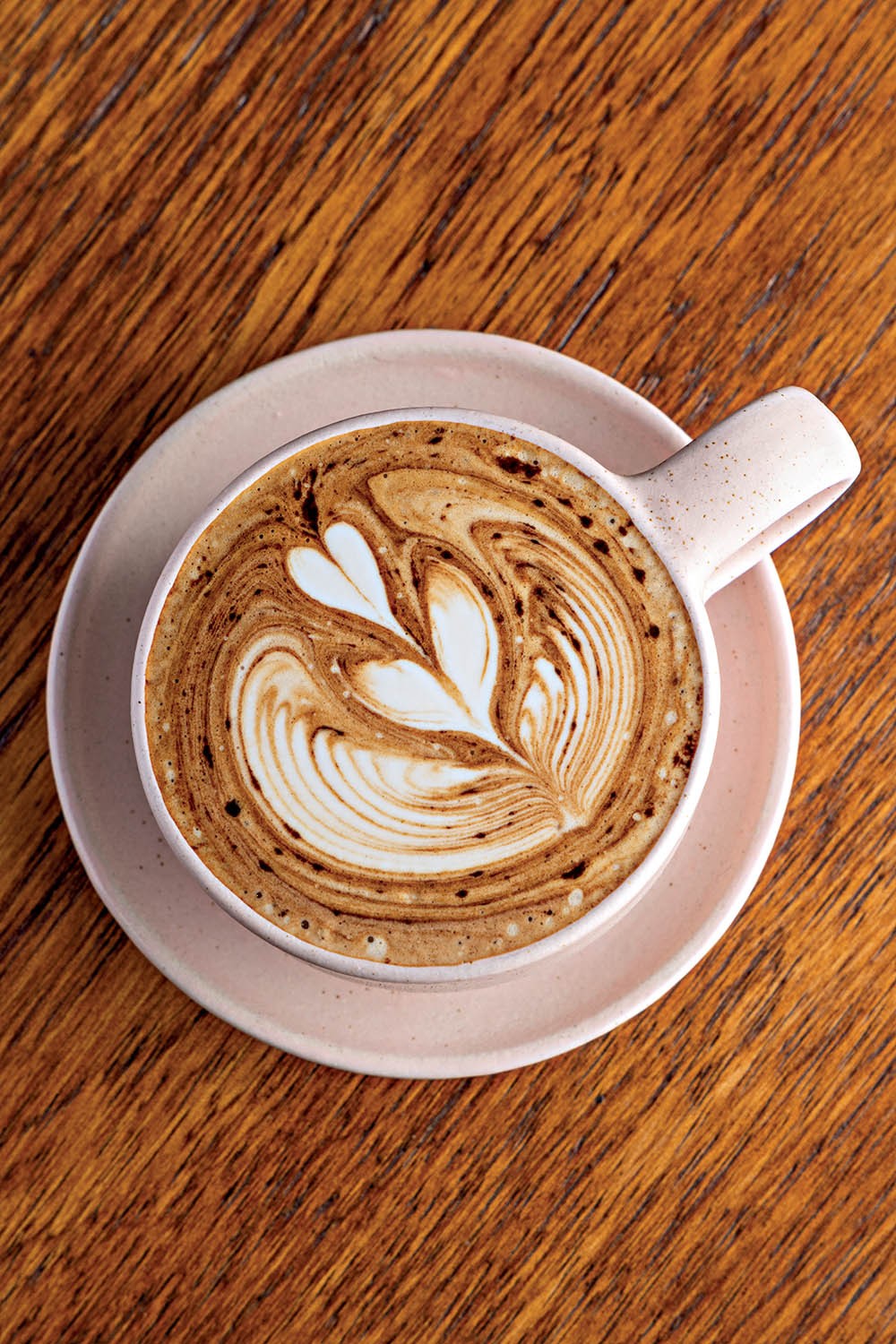 X&iacute;cara de cappuccino com arte latte de folhas e cora&ccedil;&otilde;es, em tons de marrom e branco, sobre um pires rosa claro e uma mesa de madeira escura