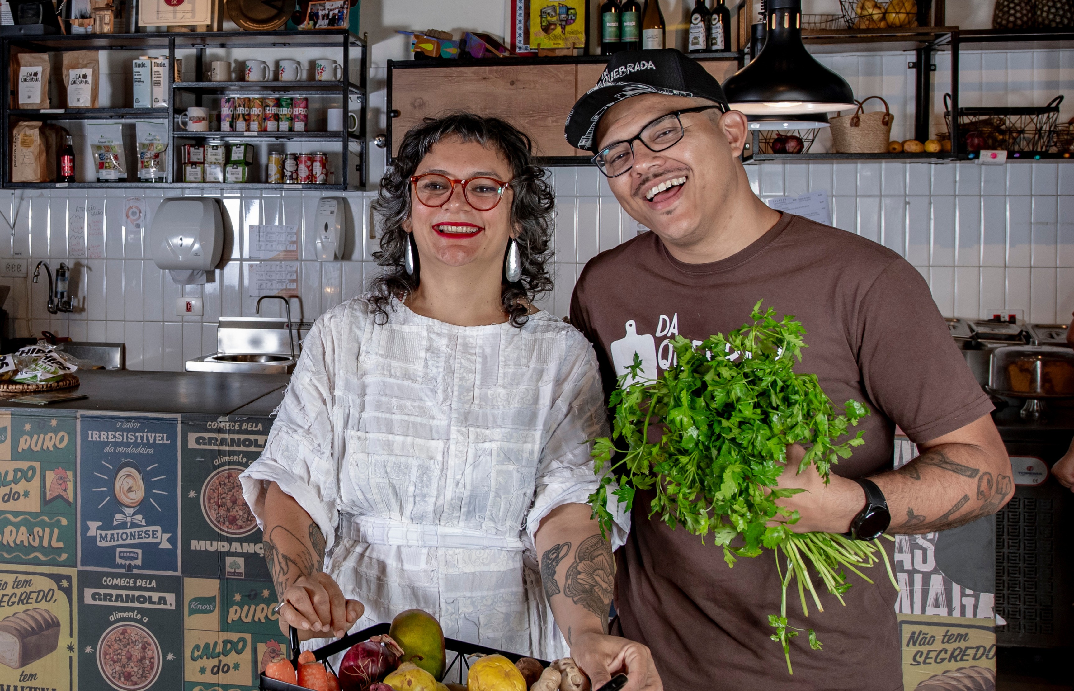 Um casal sorridente em uma cozinha. A mulher, com cabelos cacheados e &oacute;culos vermelhos, segura uma cesta de frutas. O homem, de bon&eacute; e camiseta marrom, segura um ma&ccedil;o de cheiro-verde. Prateleiras com produtos e azulejos brancos ao fundo