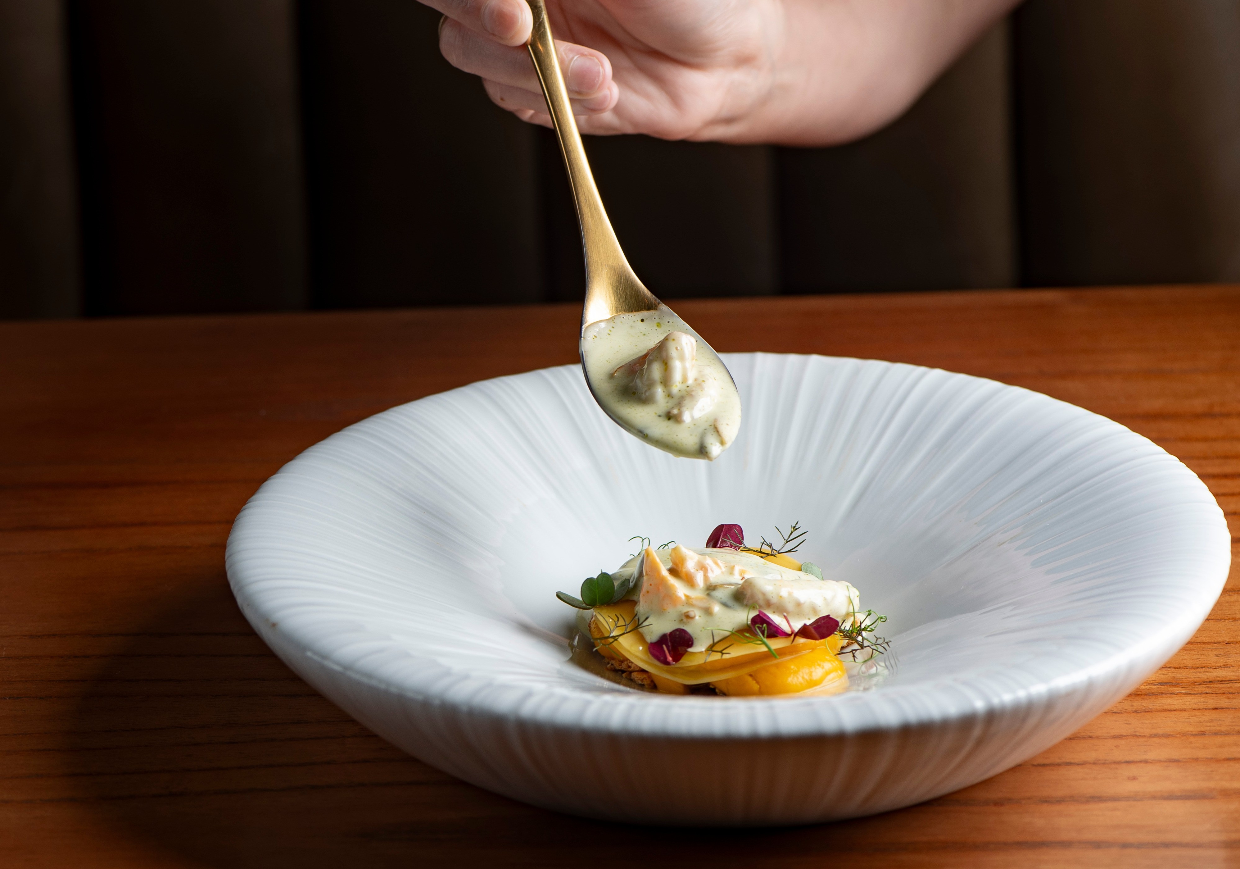 M&atilde;o de uma pessoa derramando molho cremoso de uma colher dourada sobre um prato gourmet com fatias de batata doce, peixe e folhas, em uma mesa de madeira