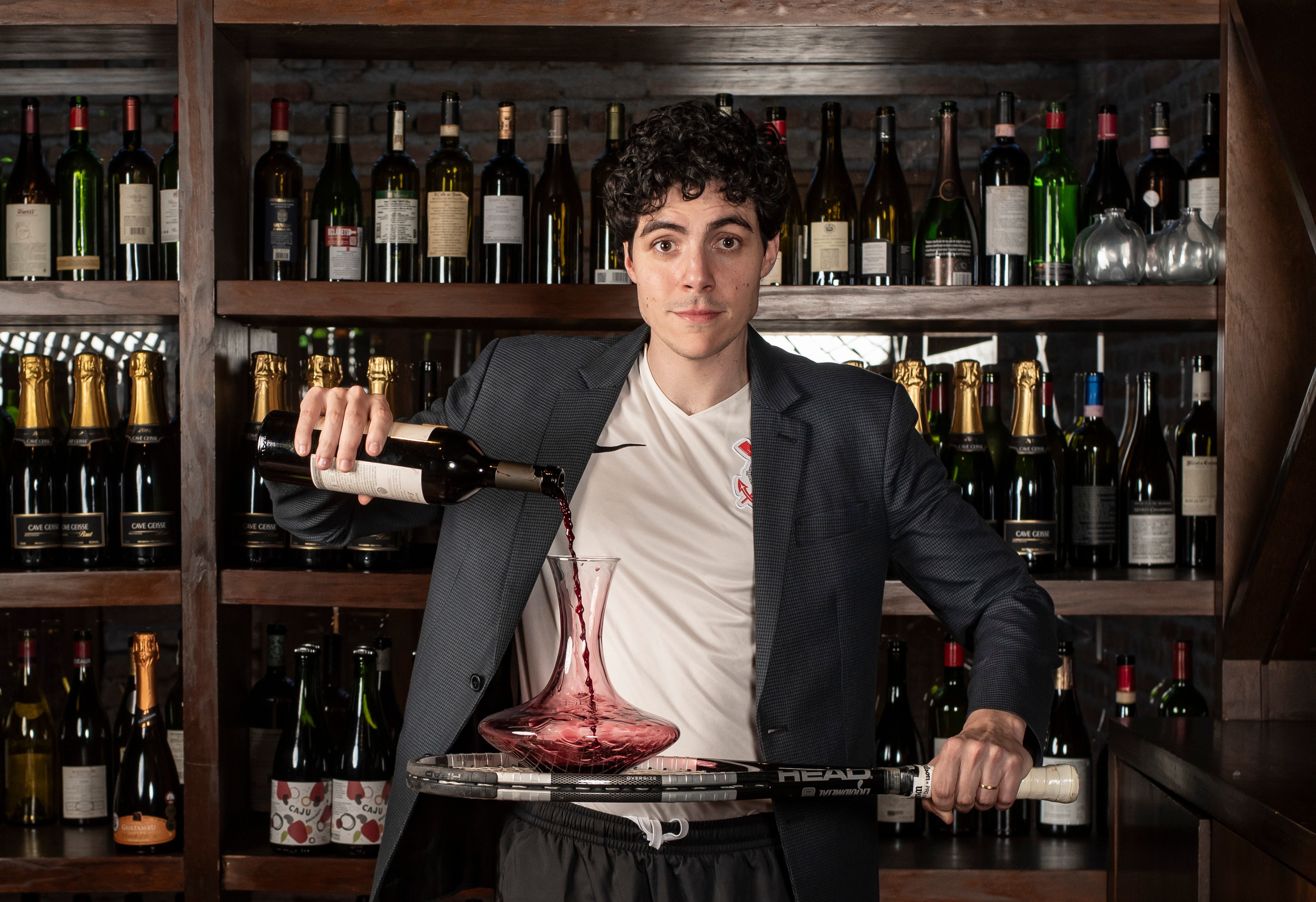 Homem jovem de cabelo cacheado escuro, vestindo blazer azul e camiseta branca, despeja vinho tinto de uma garrafa em um decanter de vidro, enquanto segura uma raquete de t&ecirc;nis. Ele est&aacute; em frente a uma prateleira de madeira com diversas garrafas de vinho e espumante.