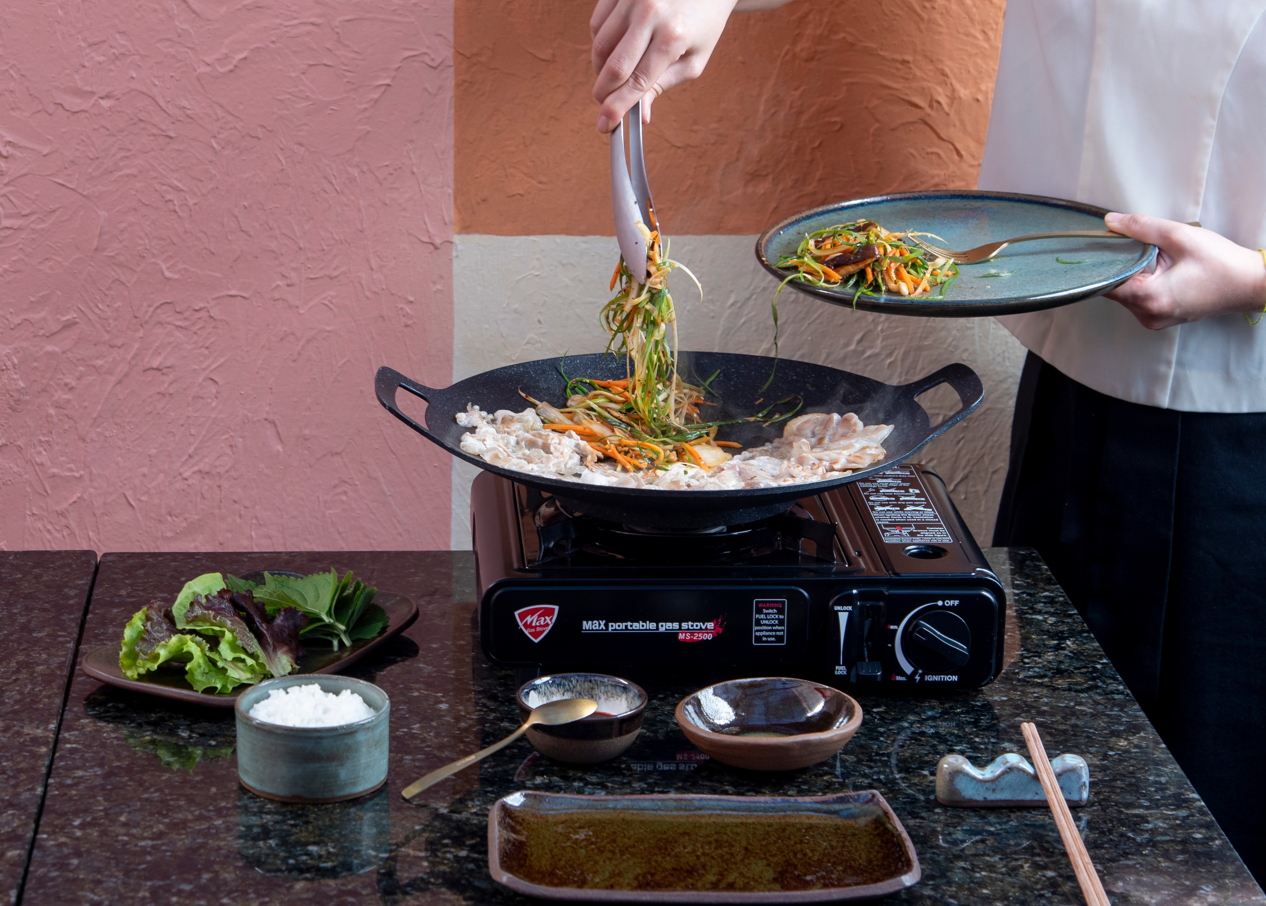 Uma pessoa com uniforme de chef branco e preto segura um prato com salada enquanto usa uma pin&ccedil;a para adicionar vegetais a uma wok com carne e legumes, sobre um fog&atilde;o port&aacute;til. Na mesa, h&aacute; um prato com folhas de alface, um pote com arroz, tigelas pequenas e hashis. O fundo &eacute; uma parede rosa e laranja