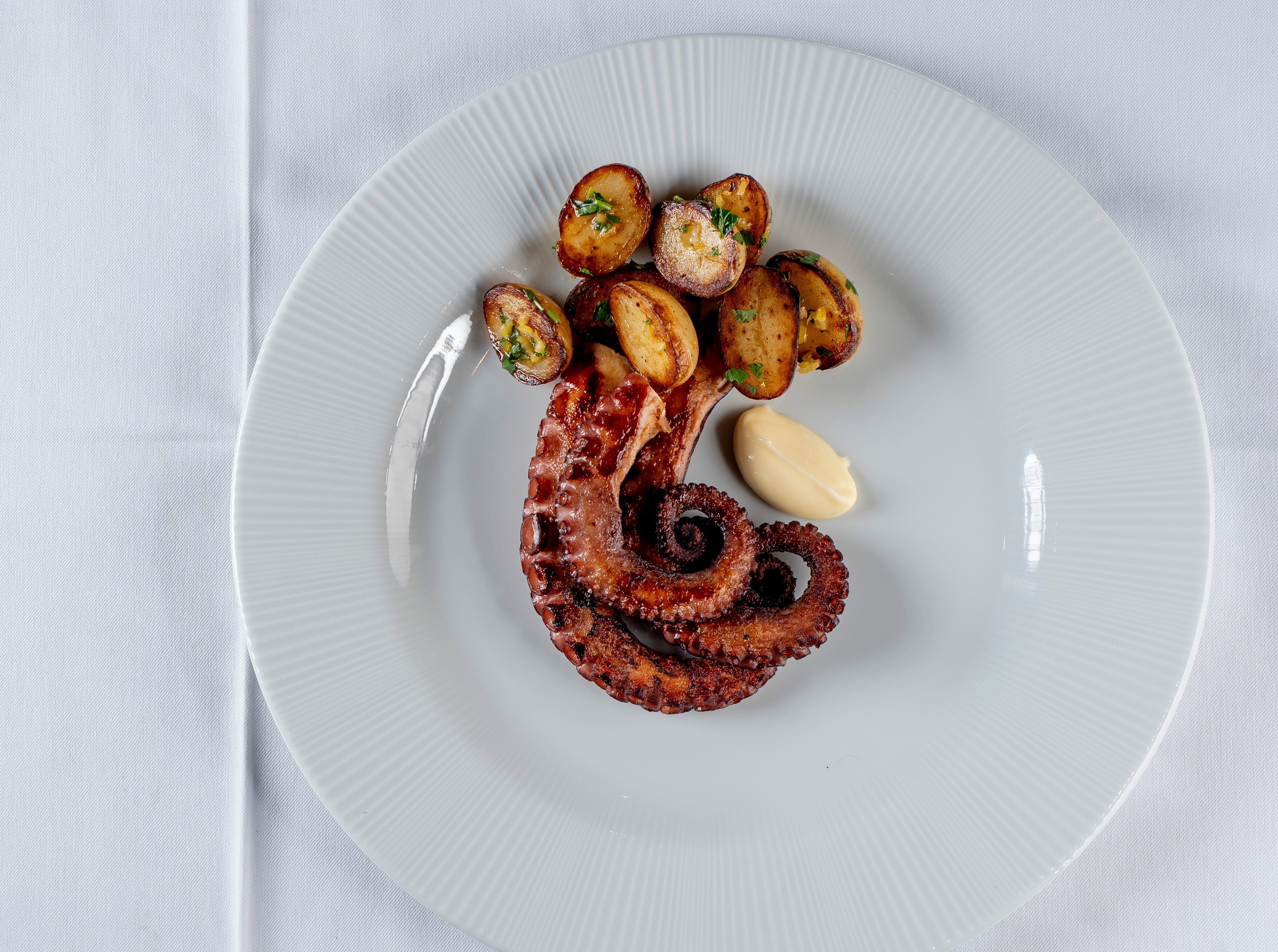 Prato de polvo grelhado com batatas assadas e um molho cremoso, servido em um prato branco sobre uma toalha de mesa branca