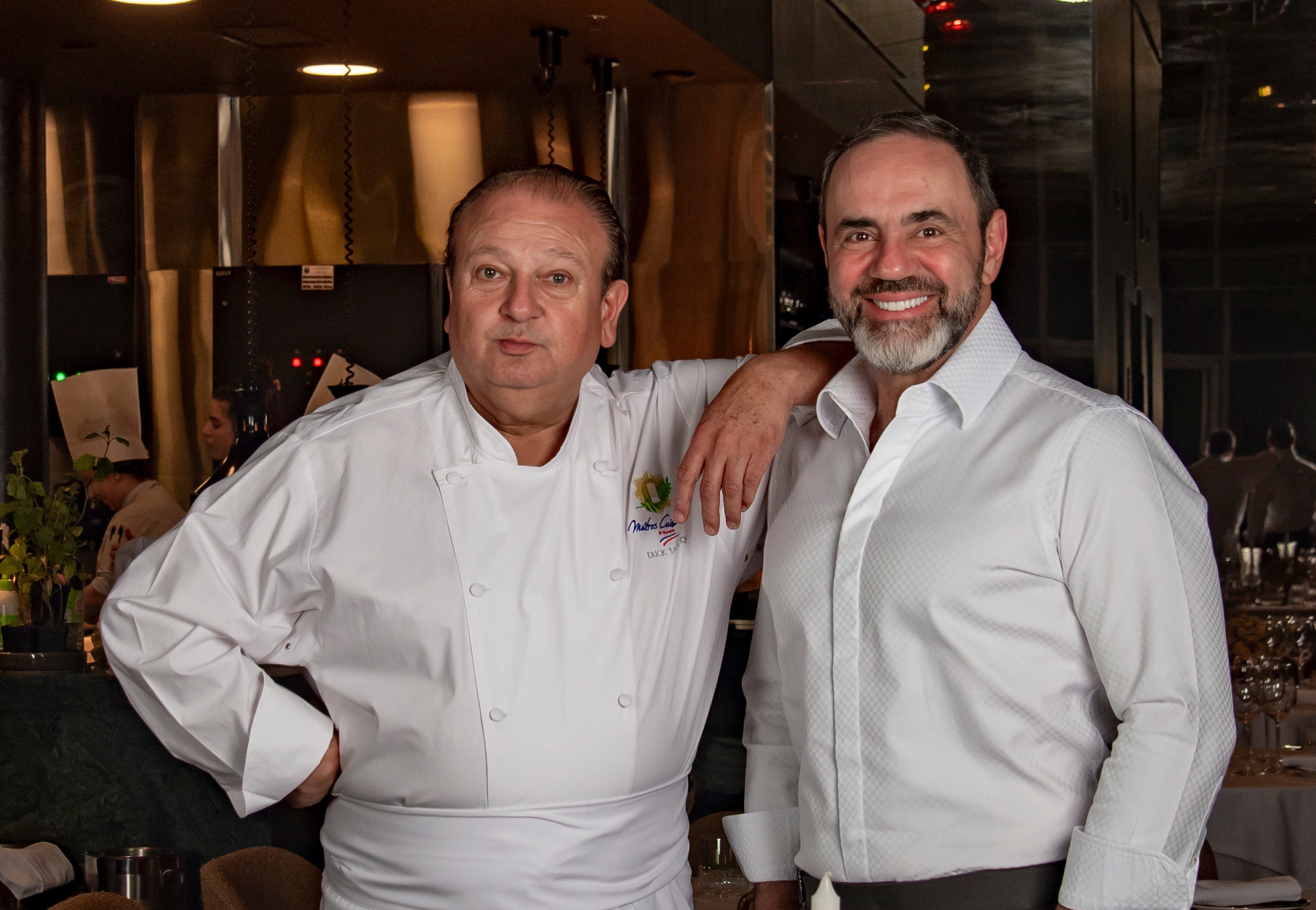 Dois homens sorridentes, um chef de cozinha e um empres&aacute;rio, posam juntos em um restaurante. O chef, &agrave; esquerda, veste d&oacute;lm&atilde; branco e avental, com a m&atilde;o na cintura. O empres&aacute;rio, &agrave; direita, usa camisa social branca e tem barba grisalha, com o bra&ccedil;o apoiado no ombro do chef